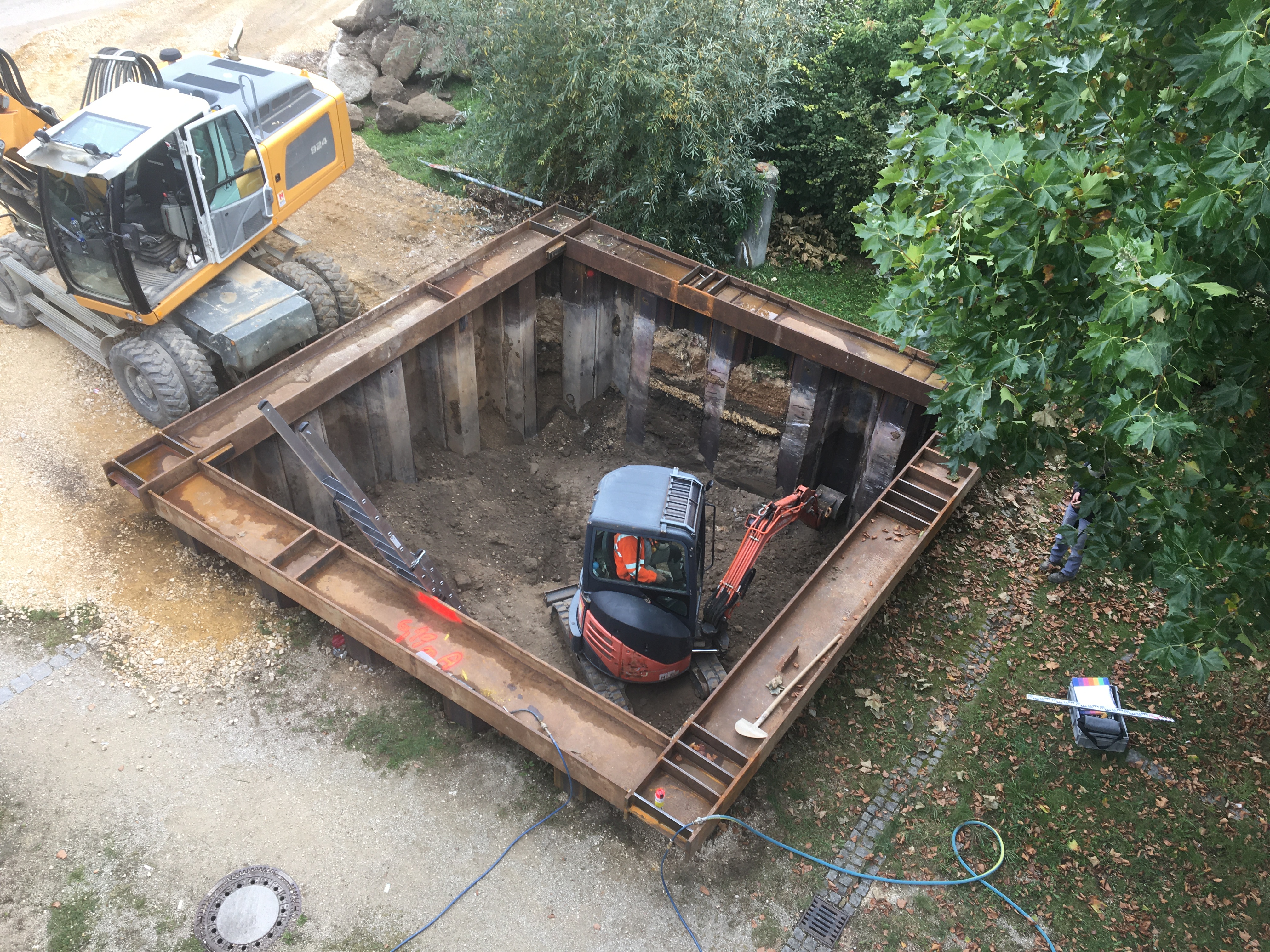 Eine quadratische Baugrube mit Spundwänden, daran lehnt eine Aluminiumleiter. Darin ein Bauarbeiter in einem rot schwarzem Minibagger zum ausbaggern der Grube. Links neben der Baugrube steht ein gelber Bagger mit leerem Führerhaus. Im Vordergrund die Baustraße, an den anderen zwei Seiten stehen Bäume.