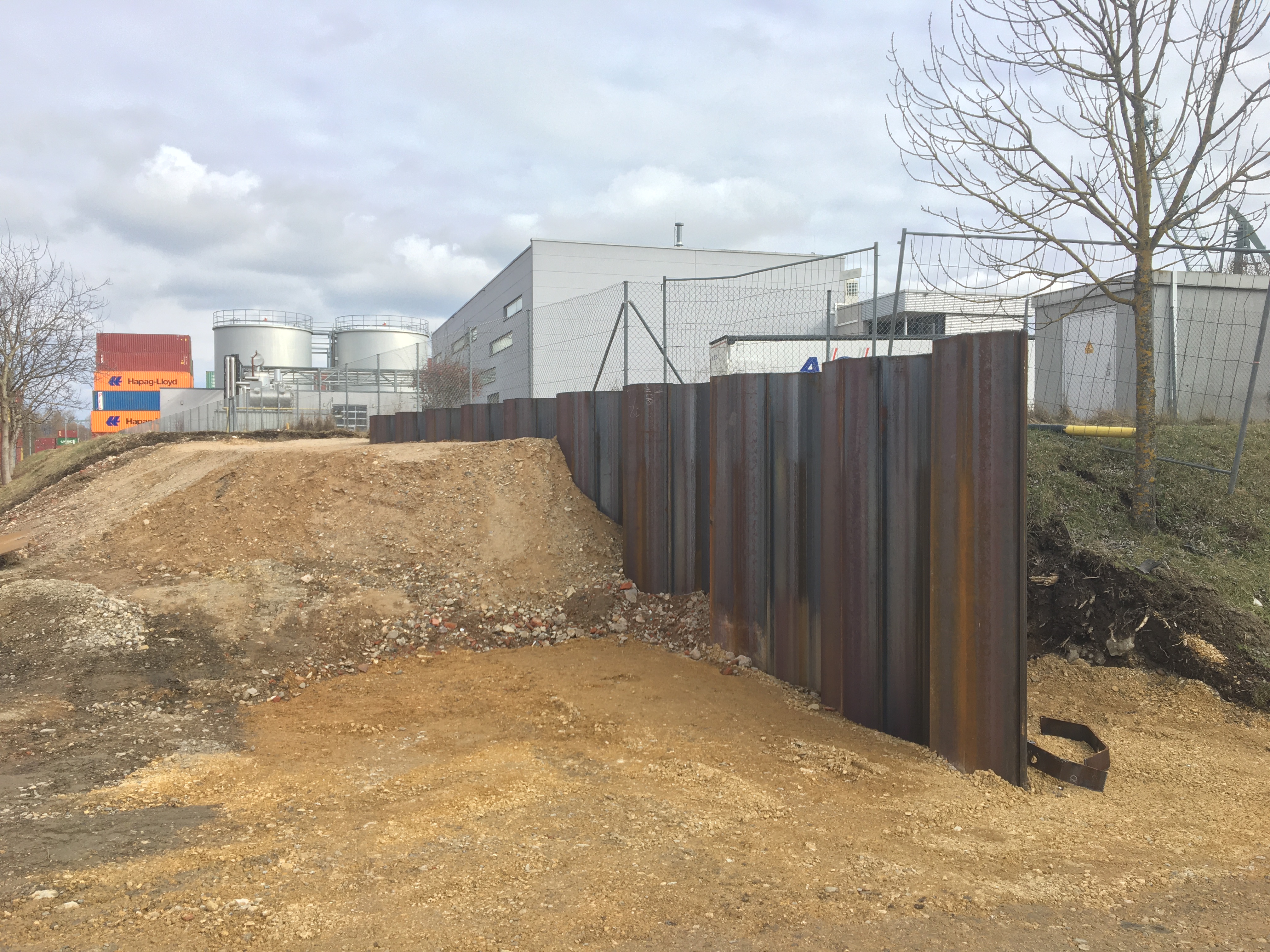 Die trapezförmige Spundwand zur Hälfte in die Böschung eingebaut. Hier endet die Hochwasserschutzmauer im Hochufer. Dahinter ein Zaun, gestapelte farbige Container, zwei Silotürme und ein Betriebsgebäude