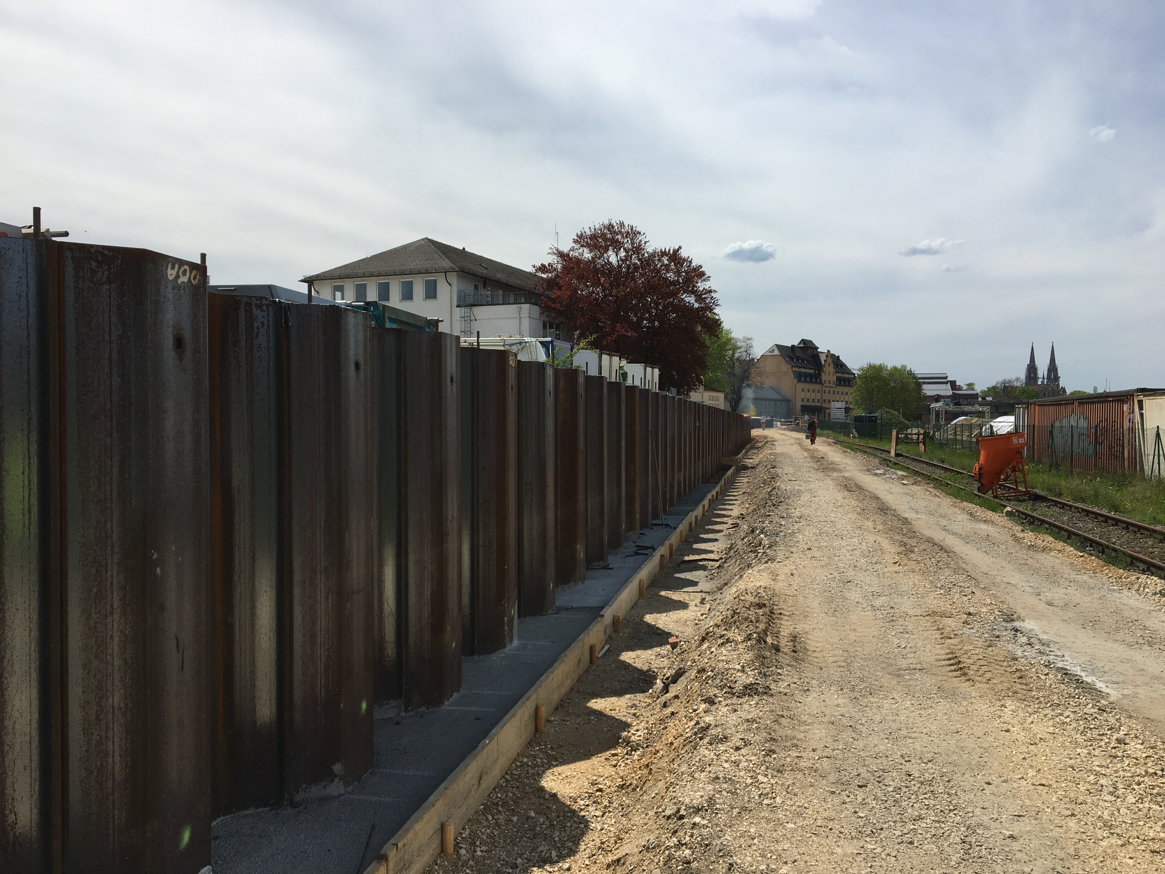 Diagonal im Bild die trapezförmige Spundwand, rechts entlang die Baustraße, auf der ein Bauarbeiter geht, ein Gleis, auf dem ein Einfülltrichter steht und ein Maschendrahtzaun. Links hinterhalb der Spundwand ein rotblättriger Baum und ein Gebäude. Im Hintergrund der IT Speicher und die Domspitzen