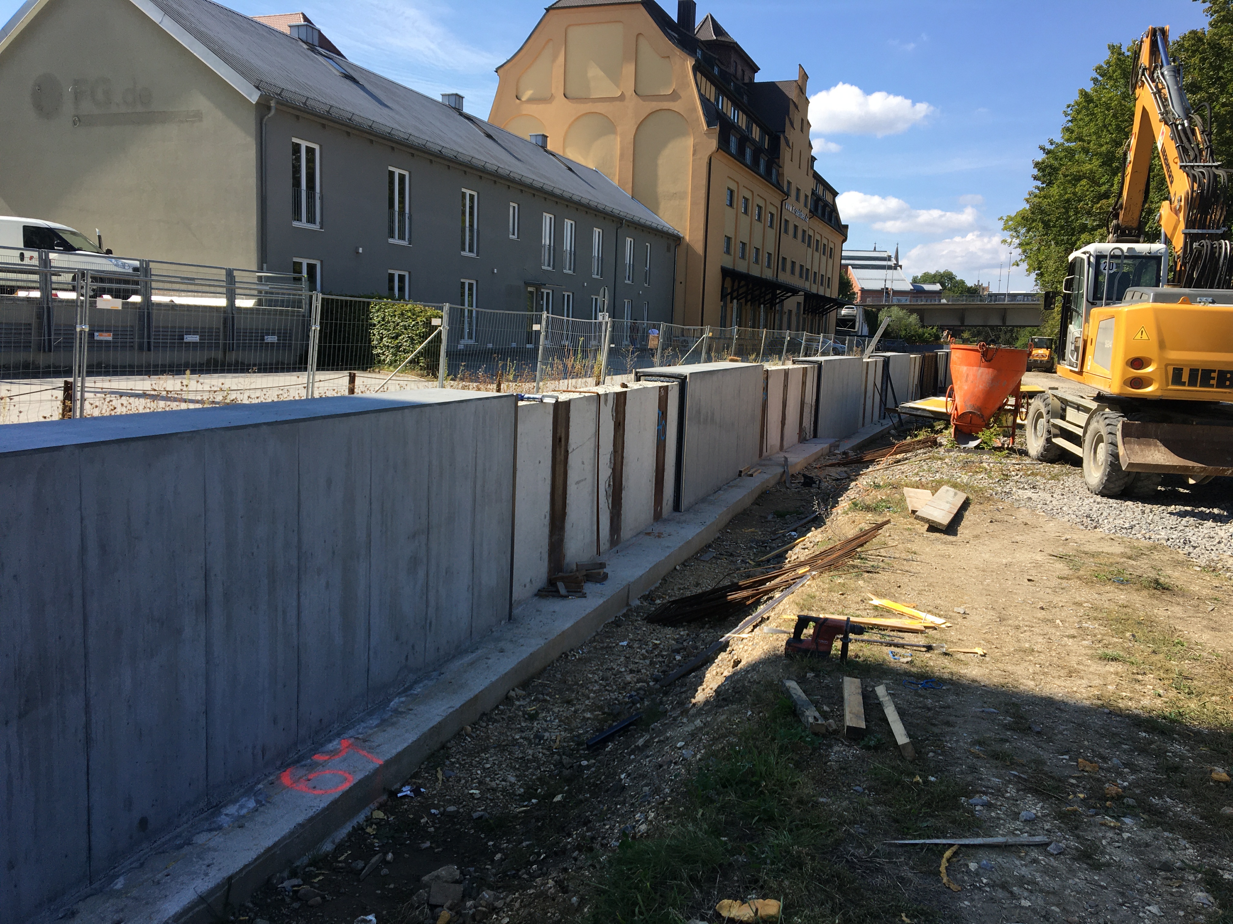 Diagonal im Bild die Hochwasserschutzwand mit grauer Betonaussenhaut, rechts davon ein Bagger, links entlang ein Bauzaun, dahinter ein graues Gebäude und der IT Speicher mit gelber Fassade. Im Hintergrund die Nibelungenbrücke