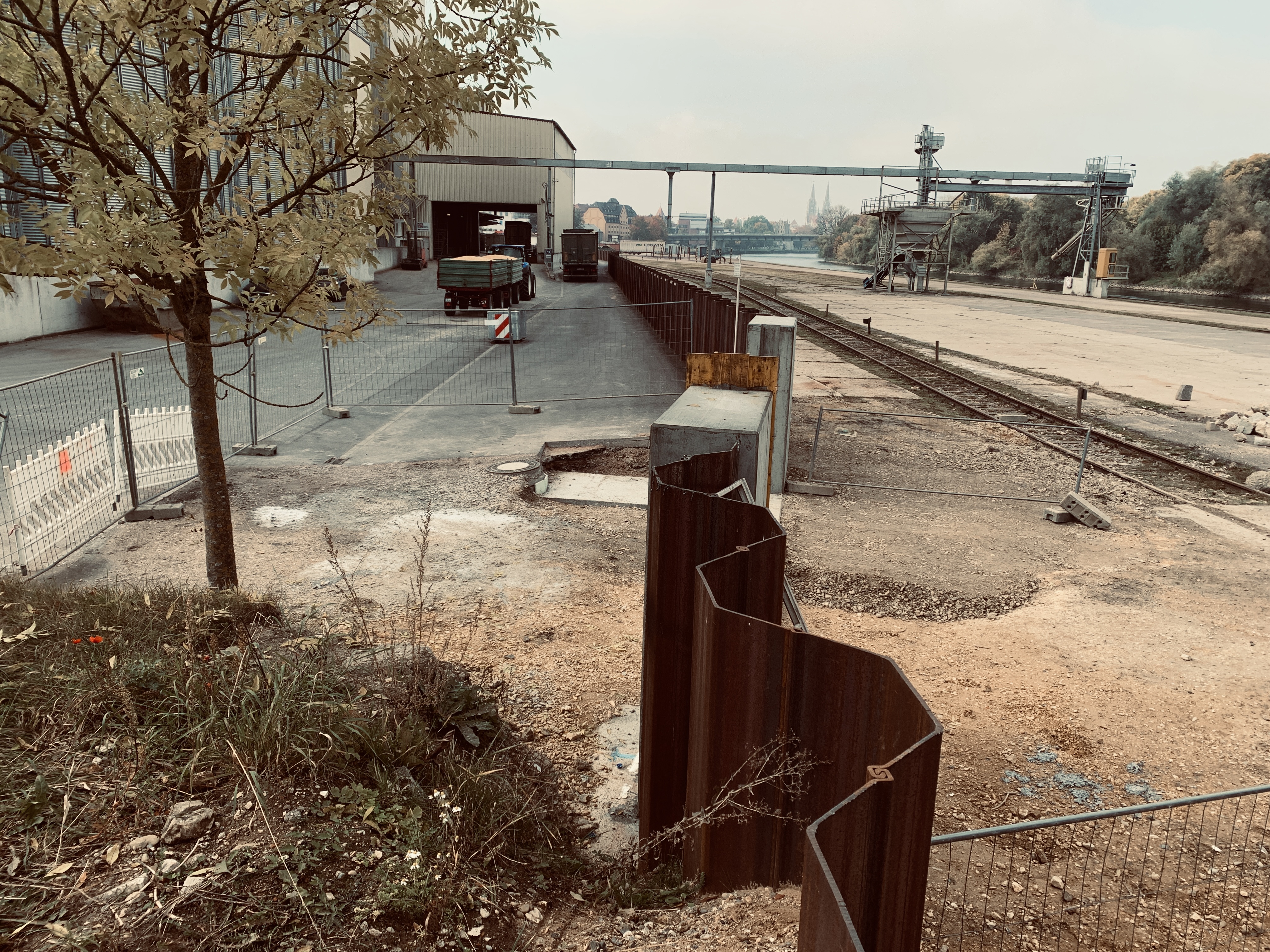 Auf dem Werksgelände Bayernhafen die eingebrachte, trapezförmige Spundwand, rechts entlang ein Gleis und eine betonierte Fläche bis zur Donau. Links im Vordergrund ein Baum, dahinter ein Bauzaun und ein Betriebsgebäude mit einem Traktor und einem LKW. Im Hintergrund sieht man unter anderem die Domspitzen und die Nibelungenbrücke