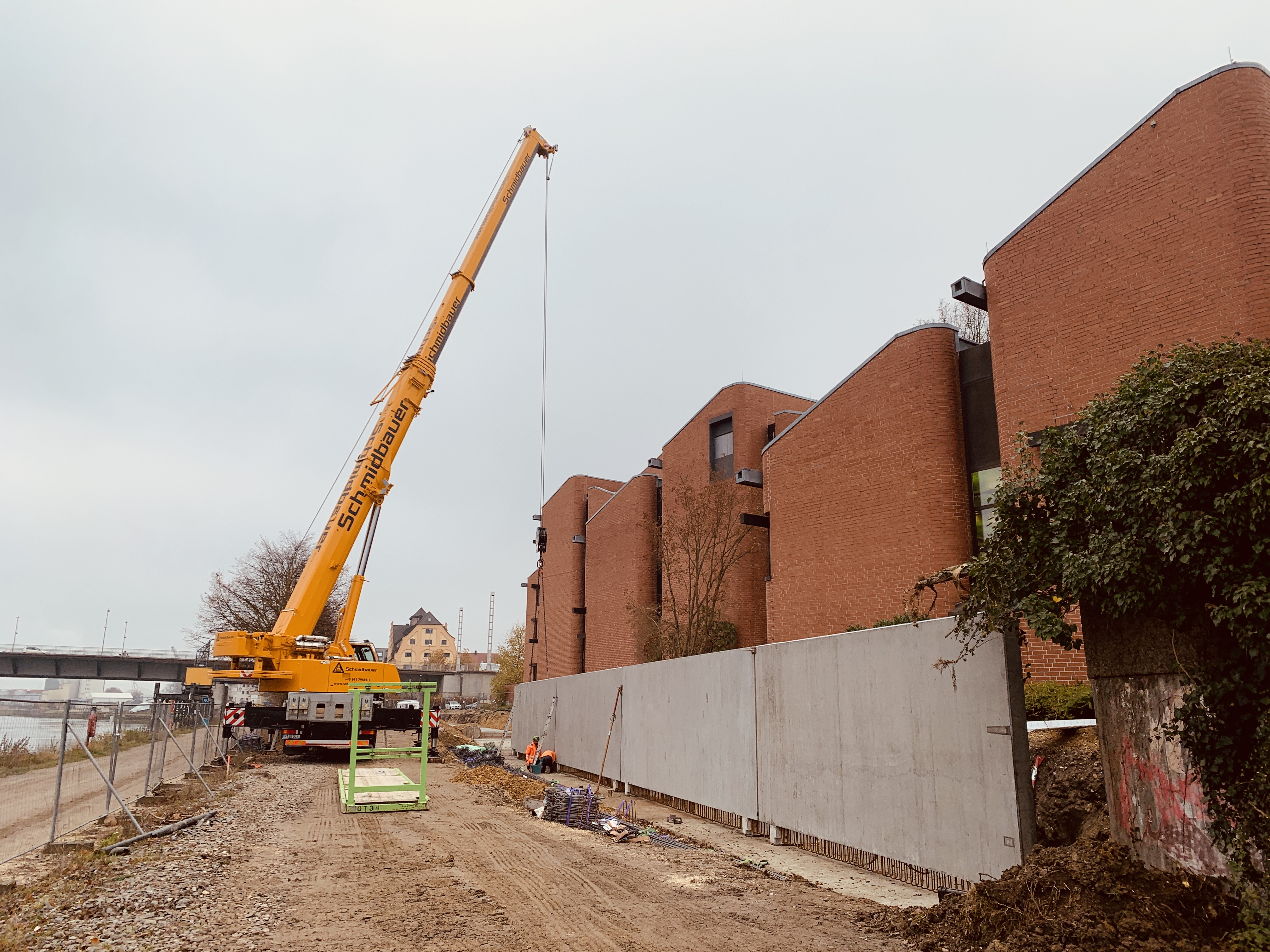 Vier aufgestellte Betonfertigteile mit zwei Bauarbeitern, rechts das AOK Gebäude mit roter Backsteinfassade, in der Mitte die Baustraße mit gelben Kran, links entlang ein Bauzaun, der Uferweg und die Donau. Im Hintergrund die Nibelungenbrücke und der IT Speicher mit gelber Fassade