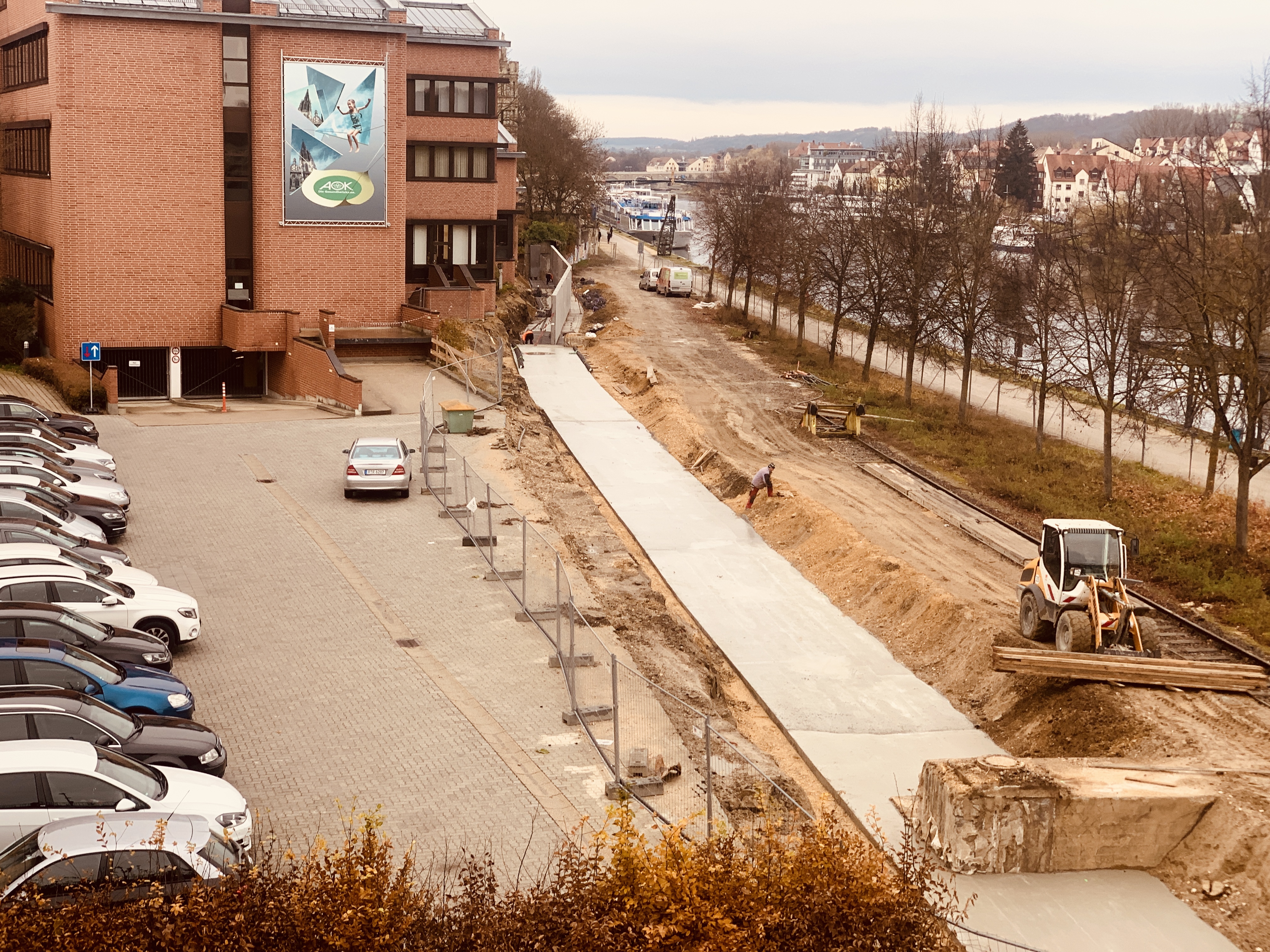 in der Mitte die Trasse für die Hochwasserschutzwand mit betonierter Sauberkeitsschicht, rechts entlang die Baustraße mit einem Baufahrzeug, eine Baumreihe, der Uferweg, die Donau und das nördliche Donauufer mit Bebauung, links entlang ein Bauzaun, ein Parkplatz mit Autos und im Hintergrund das AOK Gebäude mit roter Backsteinfassade