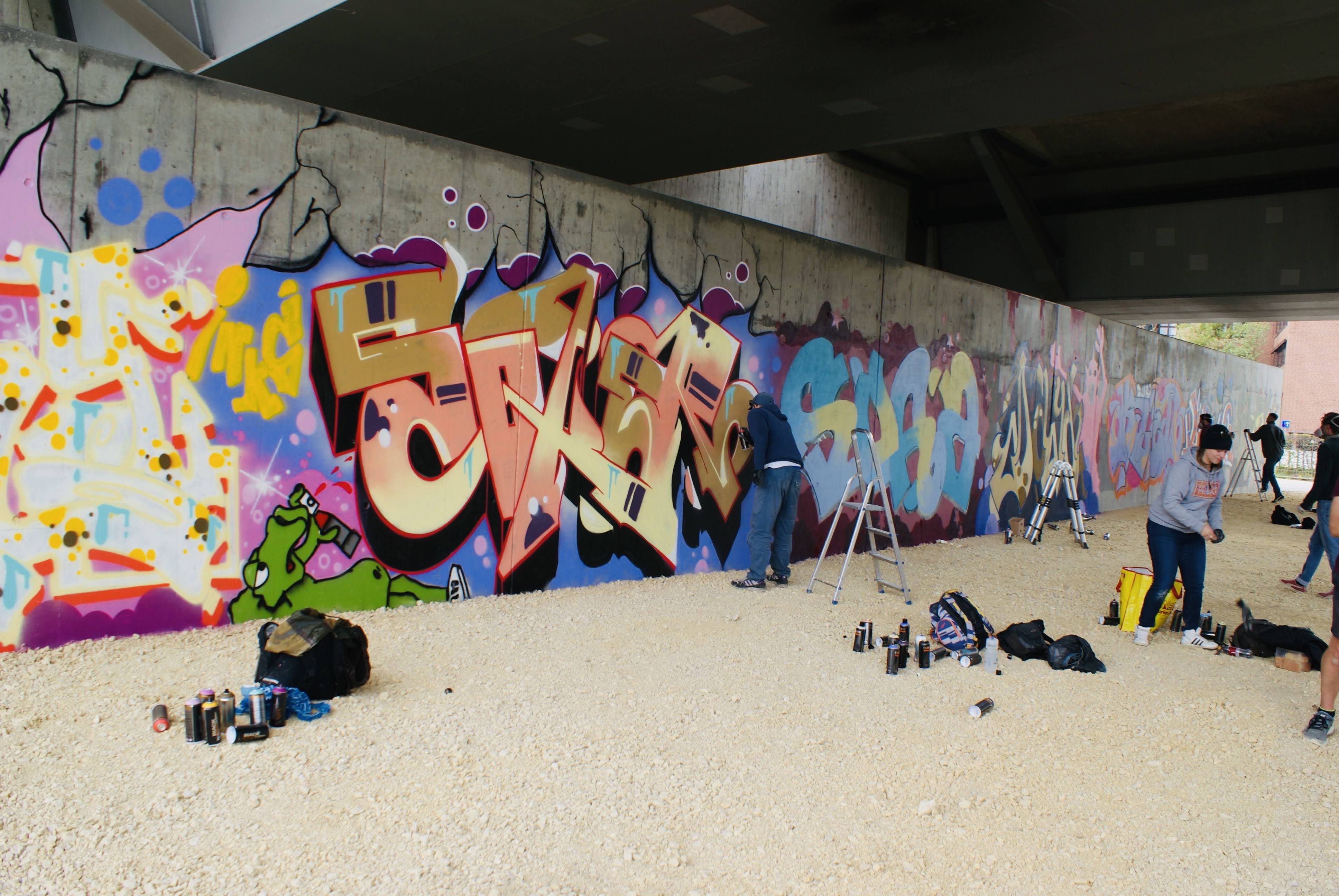 Graffiti an der HWS Wand. Die graue Betonwand mit verschieden bunten Schriftzeichen, vier Graffiti Künstler und deren Utensilien und Staffeleien.