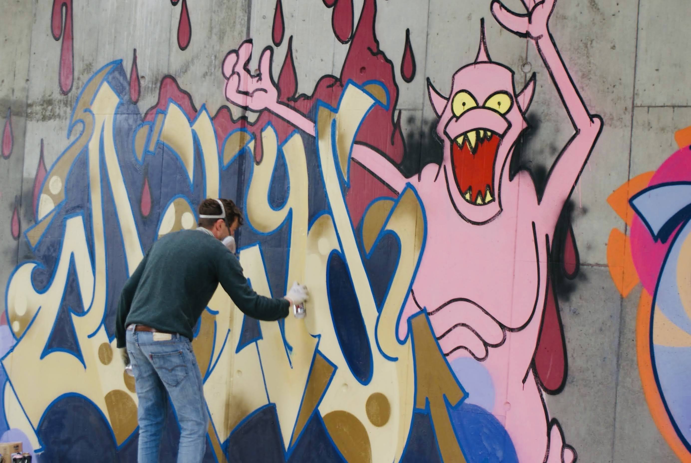 Graffiti an der HWS Wand. Rechts im Bild ein rosa Monster, welches die Arme hoch streckt. Links ein Graffiti Künstler in Jeans und dunkelgrünem Pullover sprüht einen goldgelben Schriftzug mit dunkelblauem Schatten an die graue Betonwand.