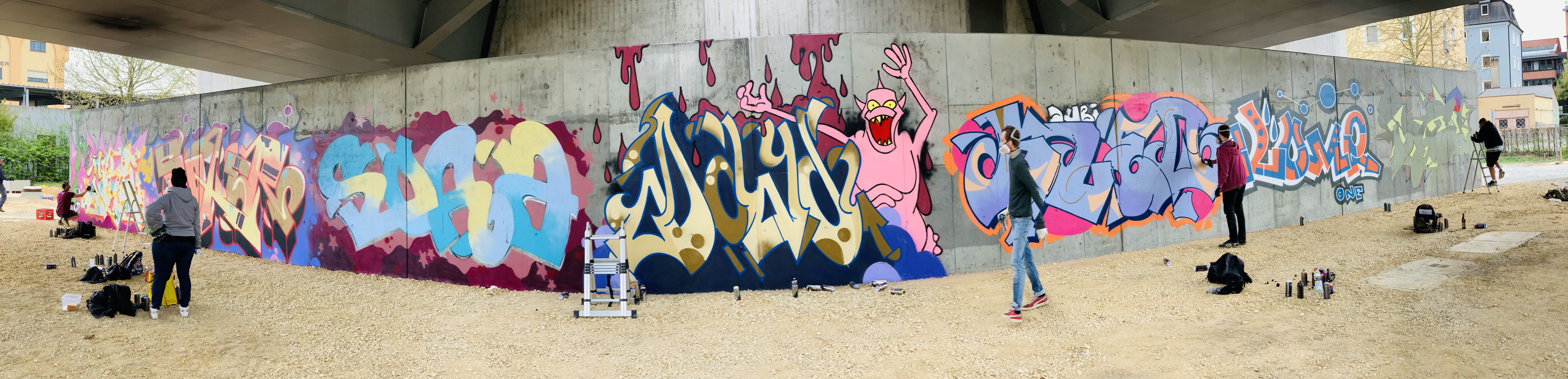 Graffiti an der HWS Wand. Die graue Betonwand in Panoramaansicht, davor sind fünf Graffitikünstler zu sehen, drei davon bearbeiten noch ihr Kunstwerk. Sieben Schriftzüge in den unterschiedlichsten Farben, in der Mitte ein rosa Monster das die Arme in die Luft streckt