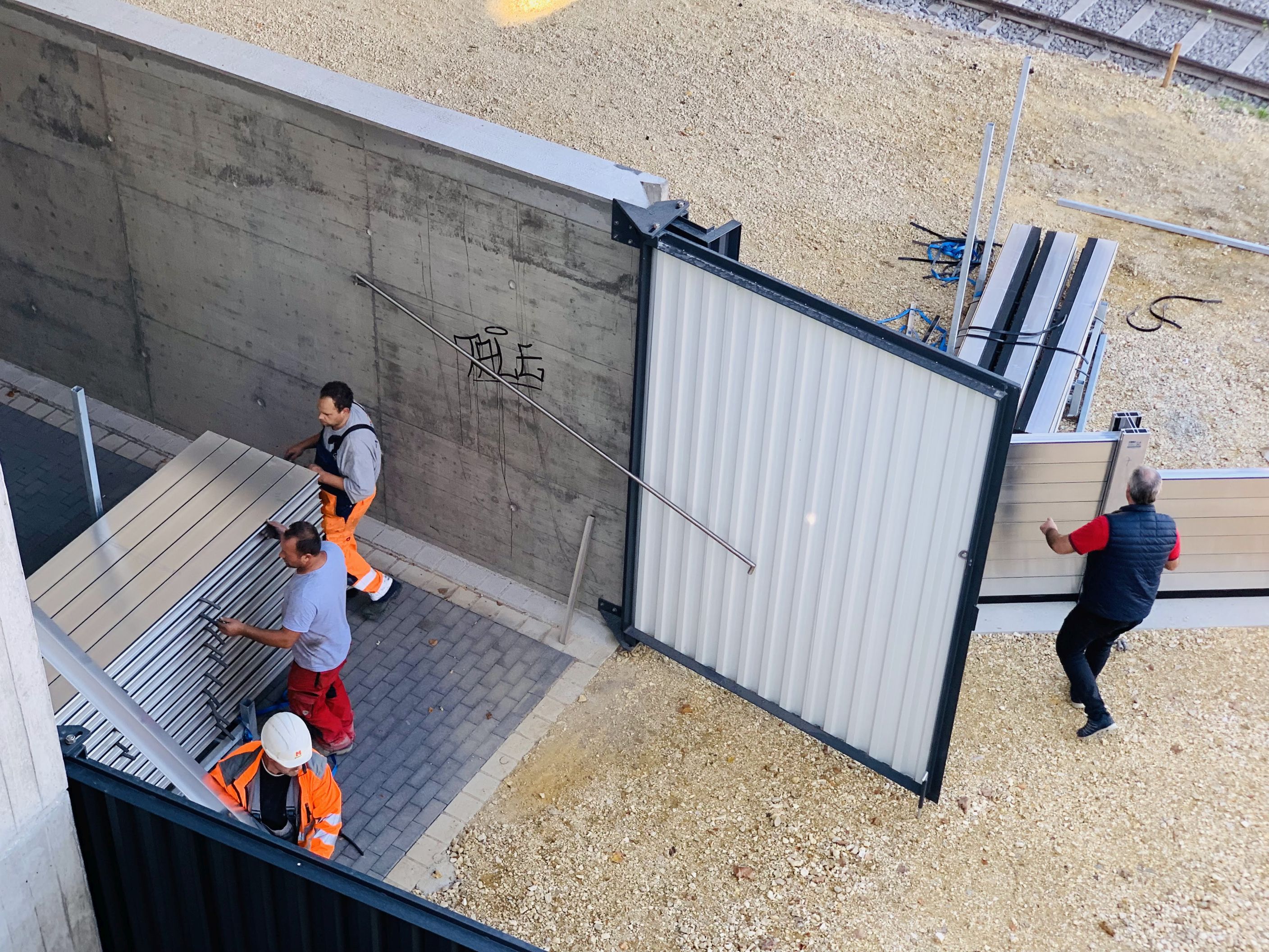 Sicht aus der Vogelperspektive in die Lagerhalle. Das Tor ist geöffnet, drei Arbeiter bringen die mobilen Elemente aus der Lagerhalle. Rechts im Bild sind schon aufgebaute mobile Elemente zu sehen. Eine Person steht davor.