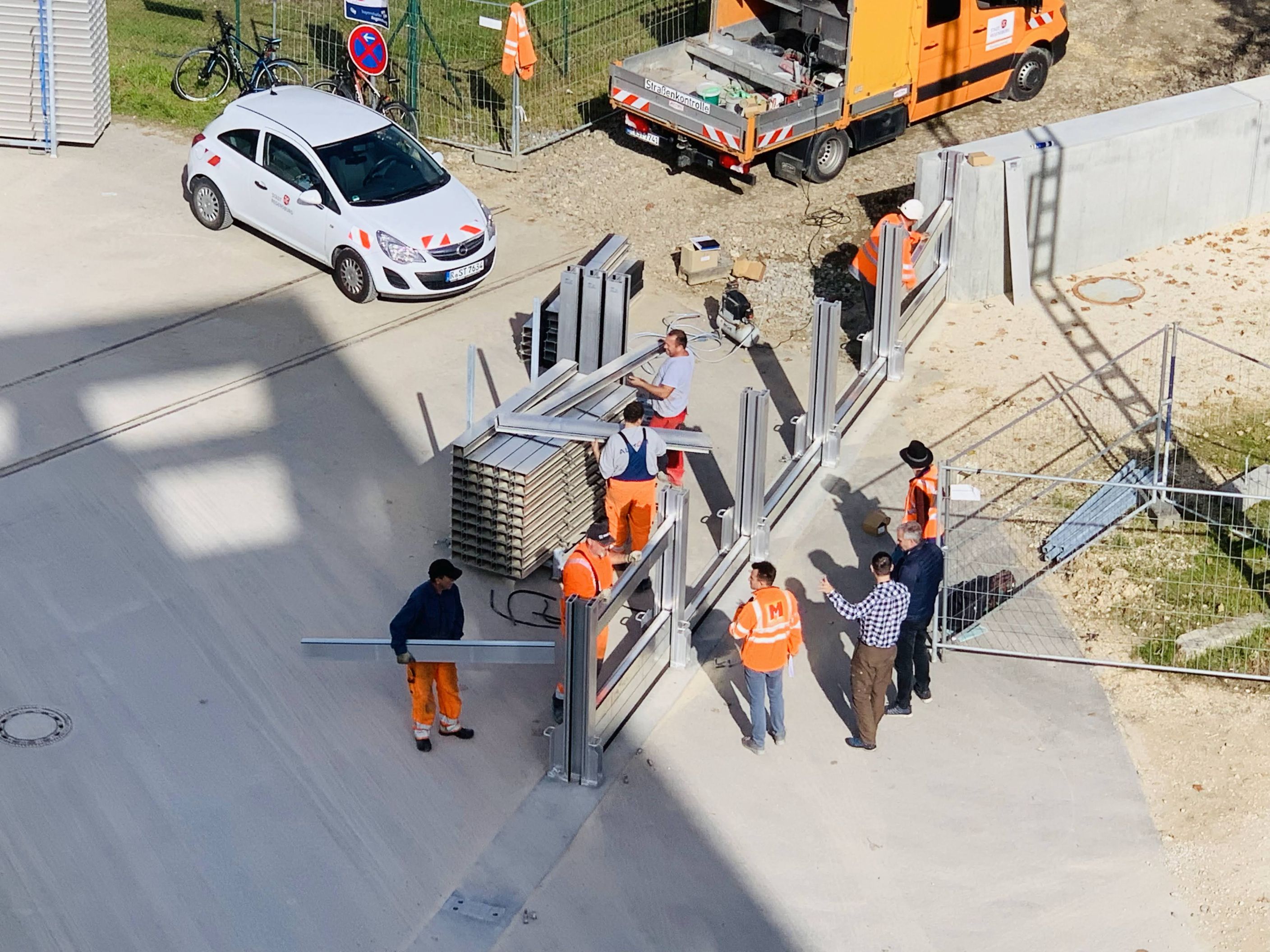 Aus der Vogelperspektive. Quer durch das Bild verläuft die Trasse zum Aufbau der mobilen Elemente, die an die Hochwasserschutzwand anschließt. Links daneben sind die Elemente abgestellt. Fünf Arbeiter sind mit dem Aufbau beschäftigt, vier weitere Personen stehen rechts daneben. Im Hintergrund stehen Fahrzeuge, Räder und ein Bauzaun.