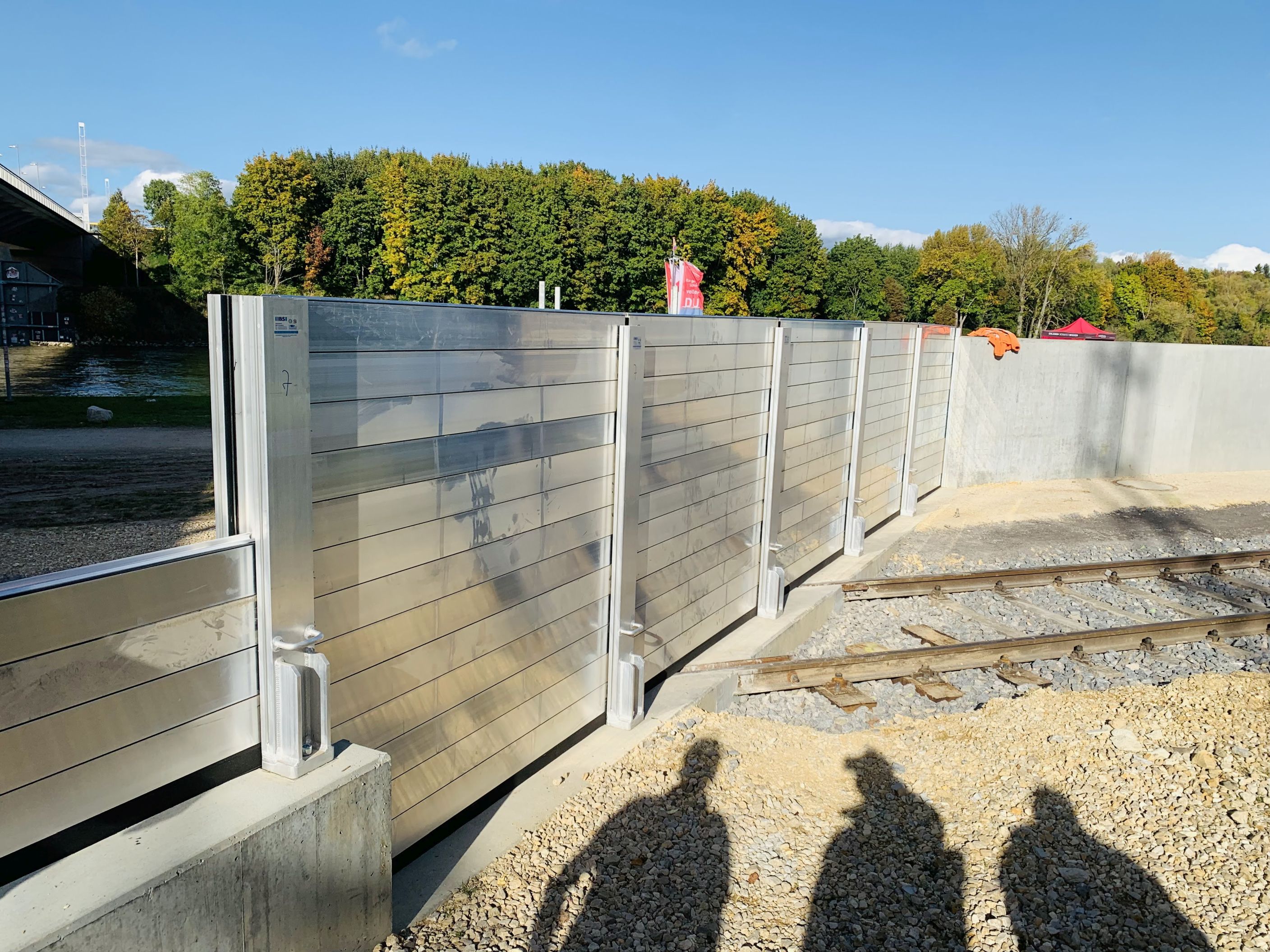 Diagonal im Bild die mobile Hochwasserschutzwand über dem Gleis, die an beiden Enden an die betonierte Hochwasserschutzwand anschließt. Rechts das Gleis, im Hintergrund Bäume , links die Nibelungenbrücke. Im Vordergrund fällt ein Schatten von drei Personen auf den geschotterten Boden.