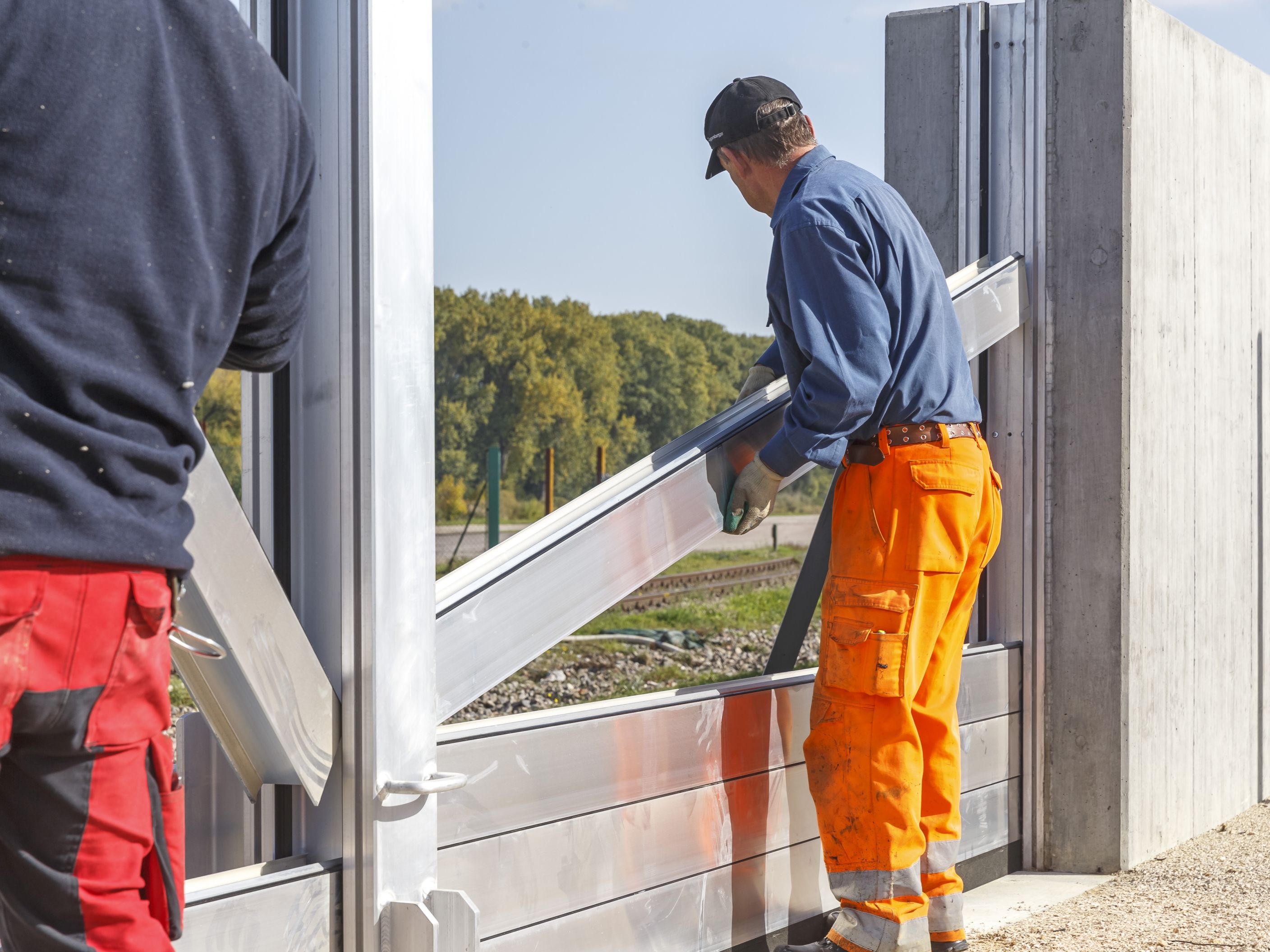 Zwei Arbeiter bauen die mobilen Elemente zwischen den Mittelsäulen und der Hochwasserschutzwand ein. Im Hintergrund queren Gleise und es stehen Bäume am Donauufer.