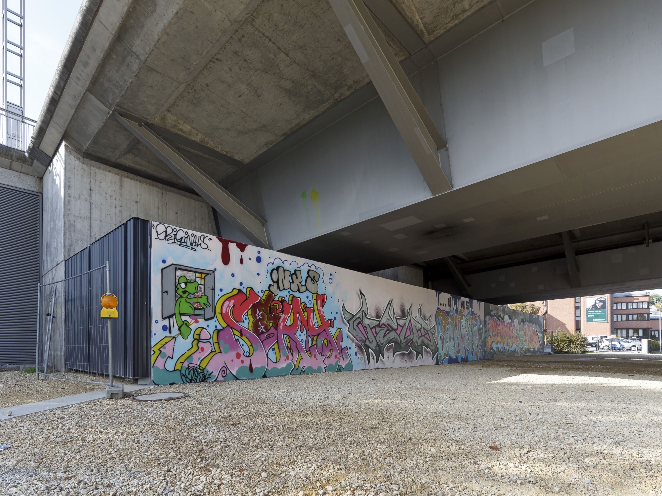 Diagonal durch das Bild verläuft die mit bunten Graffiti besprühte Hochwasserschutzwand. Am linken Ende ist das dunkelgraue Tor der Lagerhalle zu erkennen, die sich zwischen der Hochwasserschutzwand und dem Widerlager der Nibelungenbrücke befindet. Vor dem Tor steht ein Bauzaun. Der Boden vor der Hochwasserschutzwand ist geschottert, direkt über der Lagerhalle verläuft die Nibelungenbrücke.