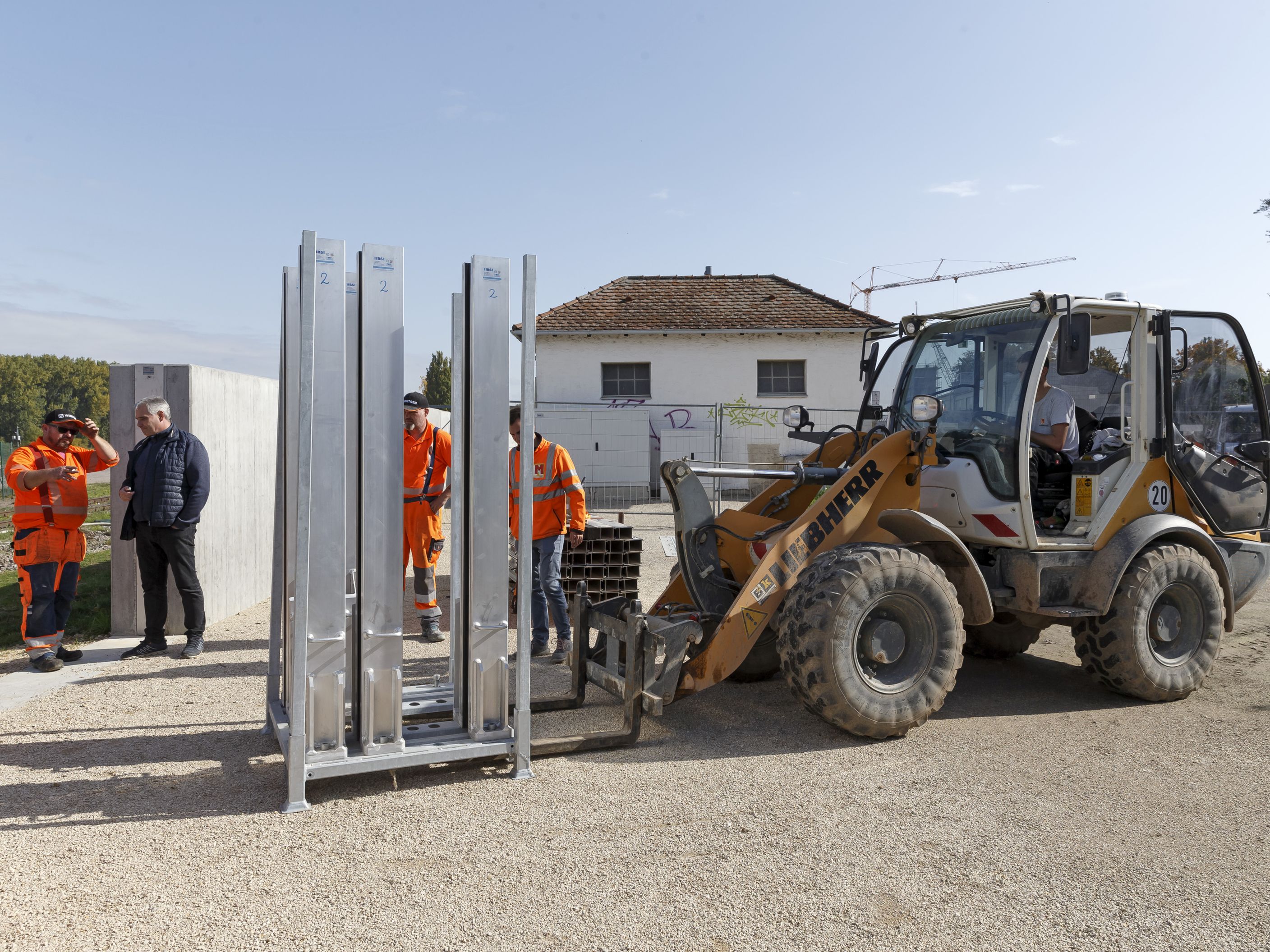 Eine Baumaschine mit Frontgabel stellt eine Palette mit Mittelsäulen für die mobilen Elemente ab, dahinter stehen Arbeiter. Links ist die Hochwasserschutzwand zu sehen, im Hintergrund Bebauung.