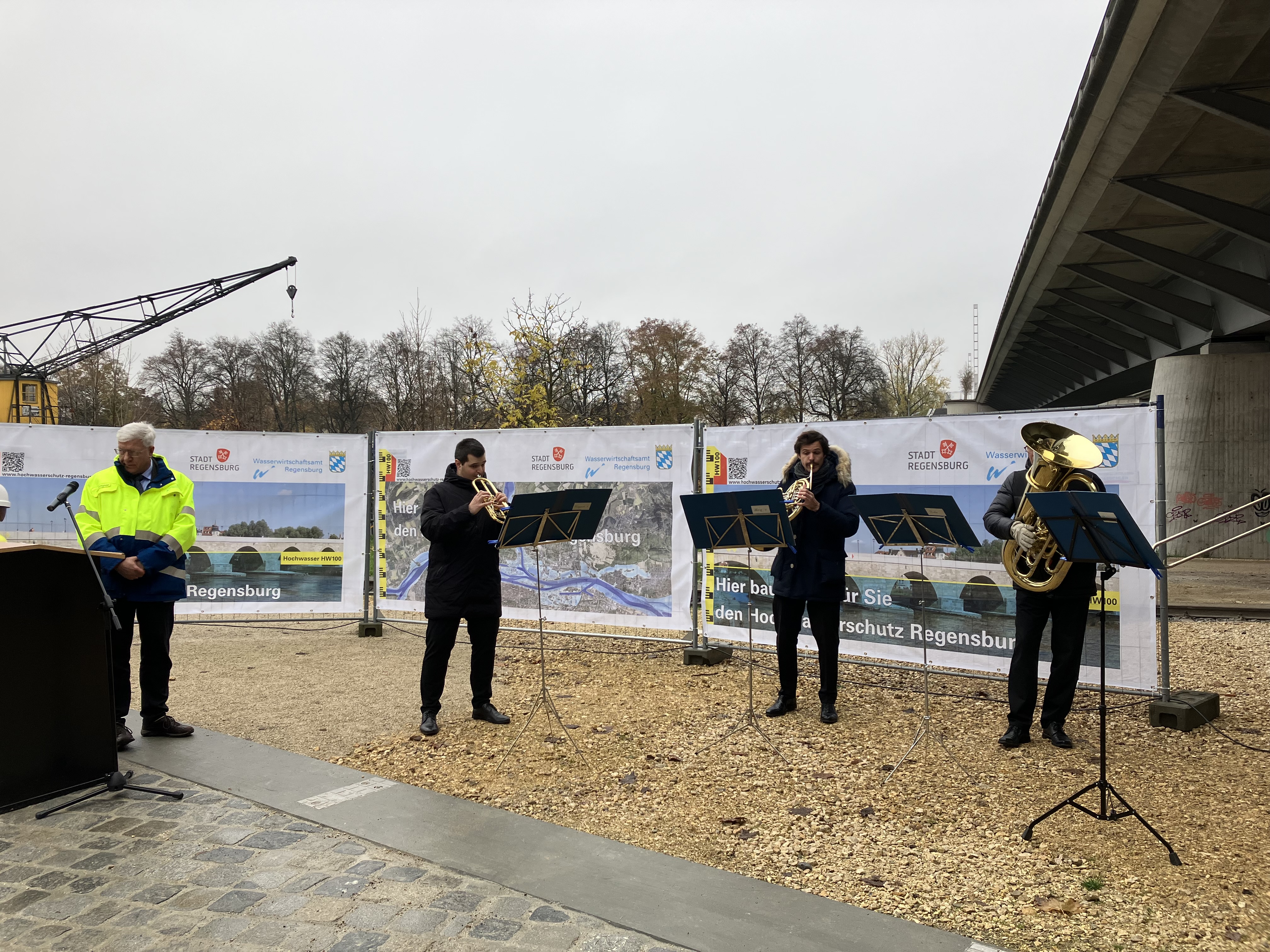 Links im Bild der Behördenleiter des WWAs, daneben drei spielende Musikanten des Philharmonischen Orchester Regensburg. Dahinter Plakate zum Hochwasserschutz Regensburg, Bäume und die Nibelungenbrücke.