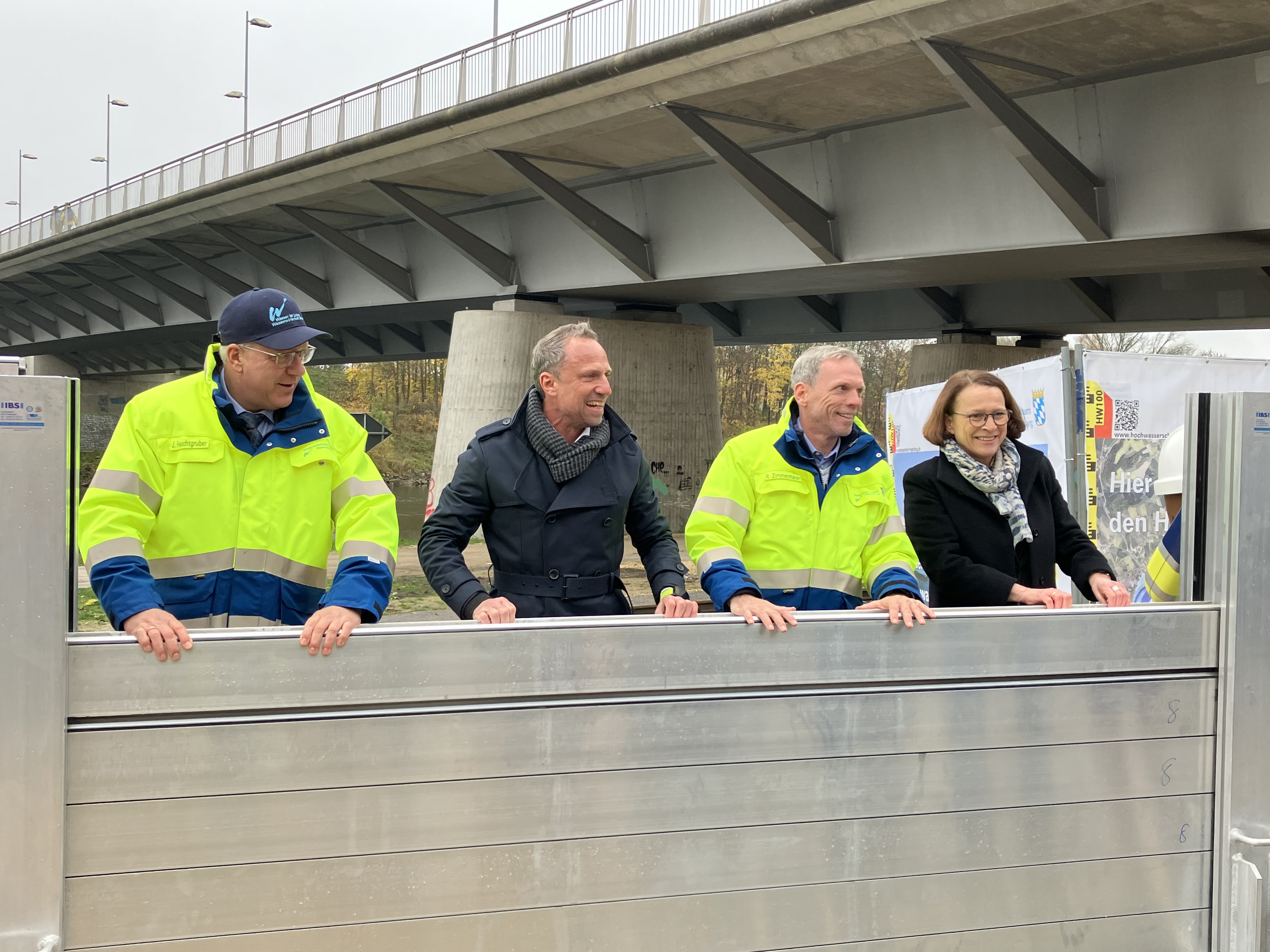 Der Amtsleiter des WWAs, der Minister, der Projektleiter des WWAs und die Oberbürgermeisterin bauen einen Dammbalken in die mobile Hochwasserschutzwand ein. Im Hintergrund diagonal im Bild die Nibelungenbrücke.