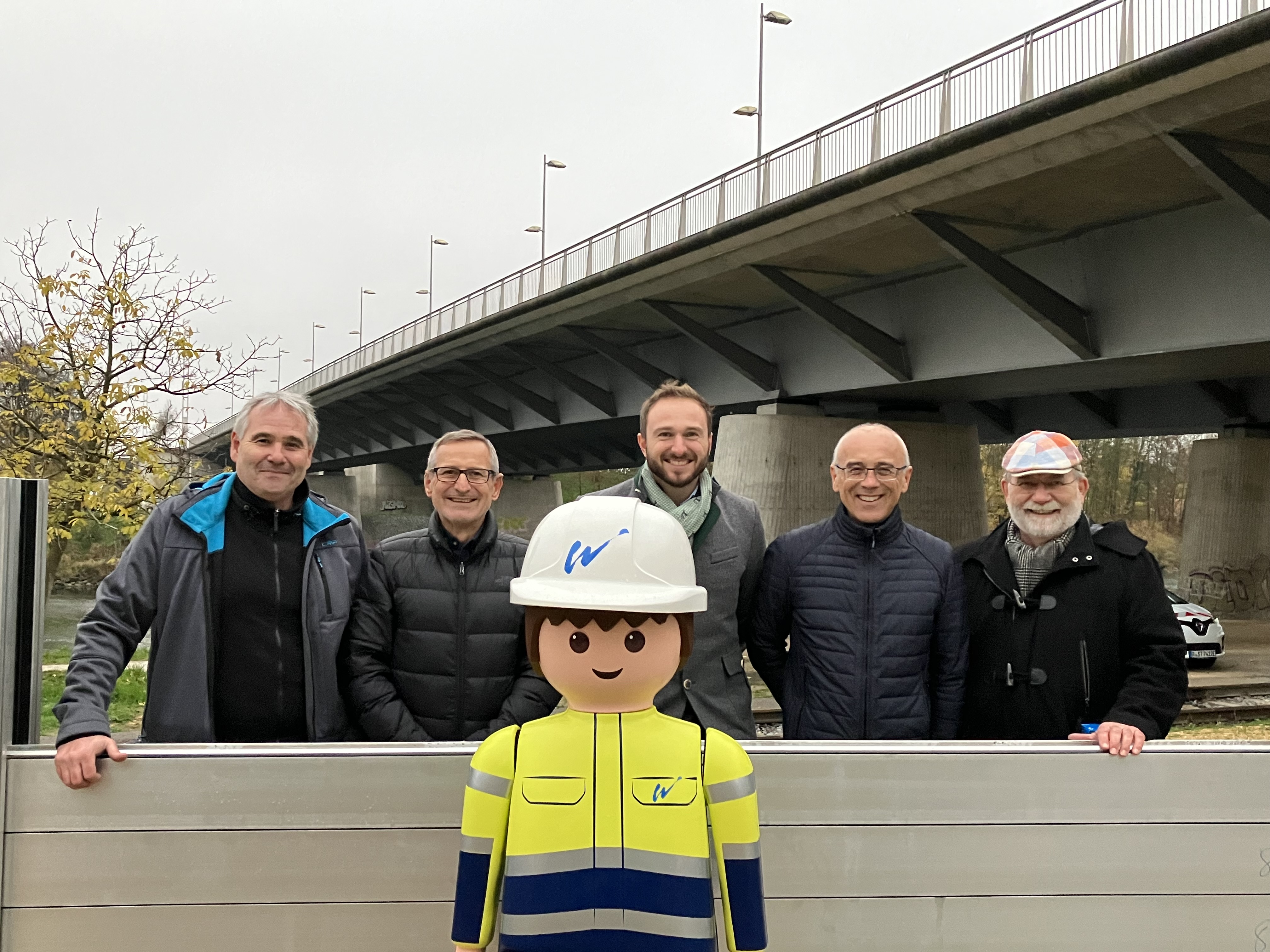 Im Vordergrund das Playmobilmännchen, dahinter die zum Teil aufgebaute mobile Hochwasserschutzwand mit fünf Mitarbeitern der Stadt Regensburg. Im Hintergrund diagonal im Bild die Nibelungenbrücke.