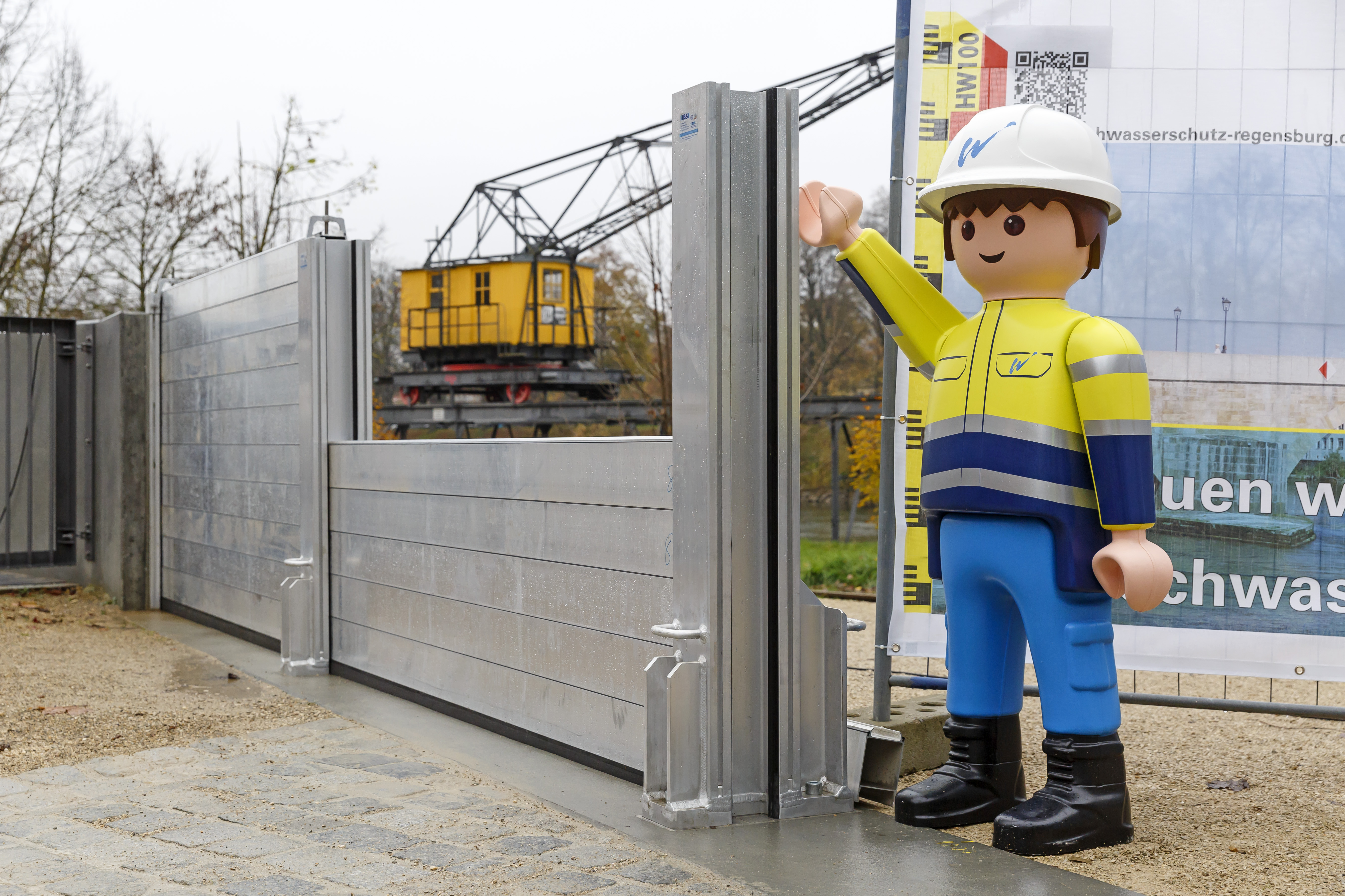 Diagonal im Bild die zur Hälfte aufgebaute mobile Hochwasserschutzwand. Dahinter ein alter gelber Hafenkran, rechts daneben ein Playmobilmännchen ca. einen Meter groß, in neongelber Wasserwirtschaftsamtschutzjacke und weißem Helm.