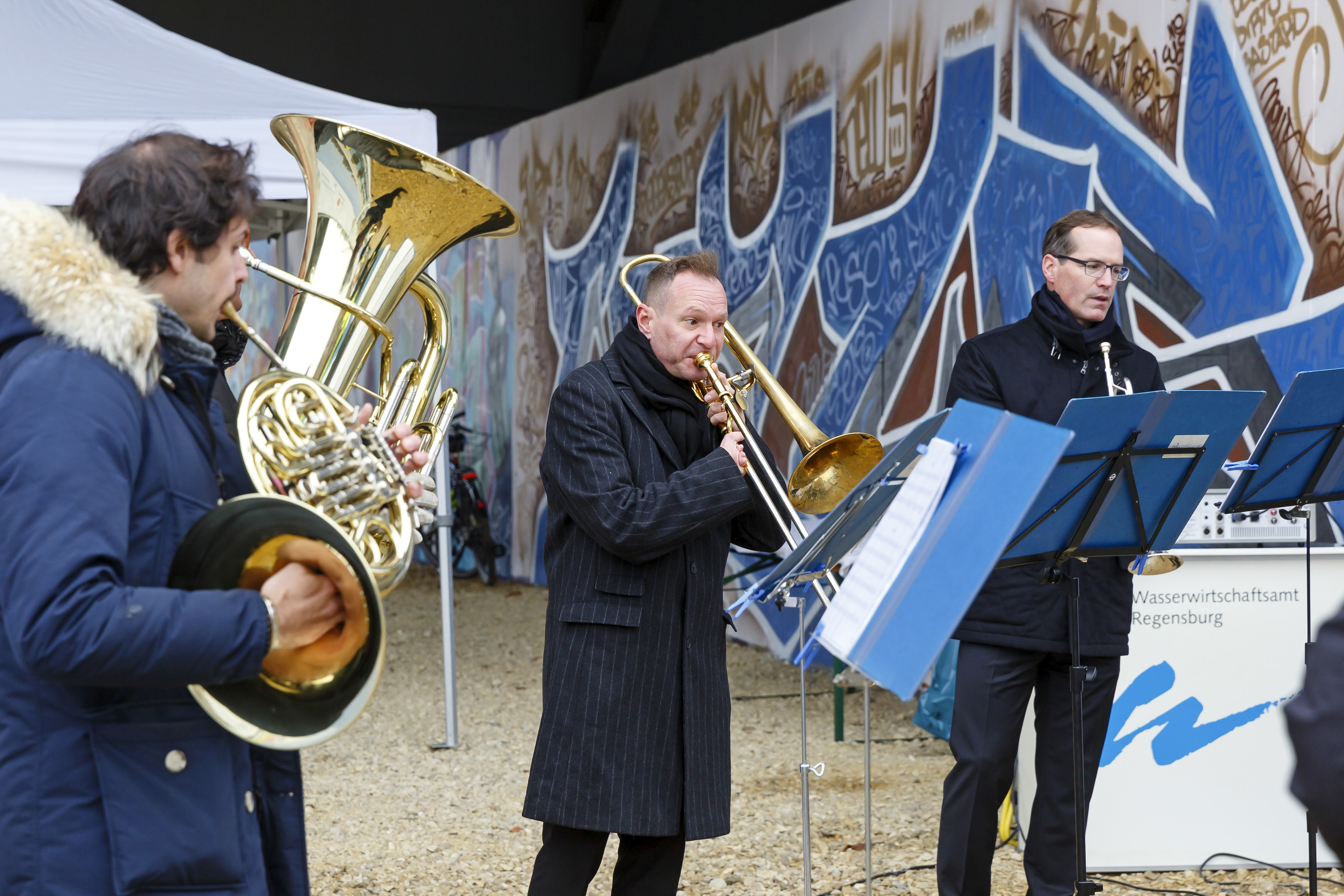 Drei Musikanten des Philharmonischen Orchester Regensburg mit Blechblasinstrumenten und Notenständern. Im Hintergrund das mit Graffiti besprühte Widerlager der Nibelungenbrücke.