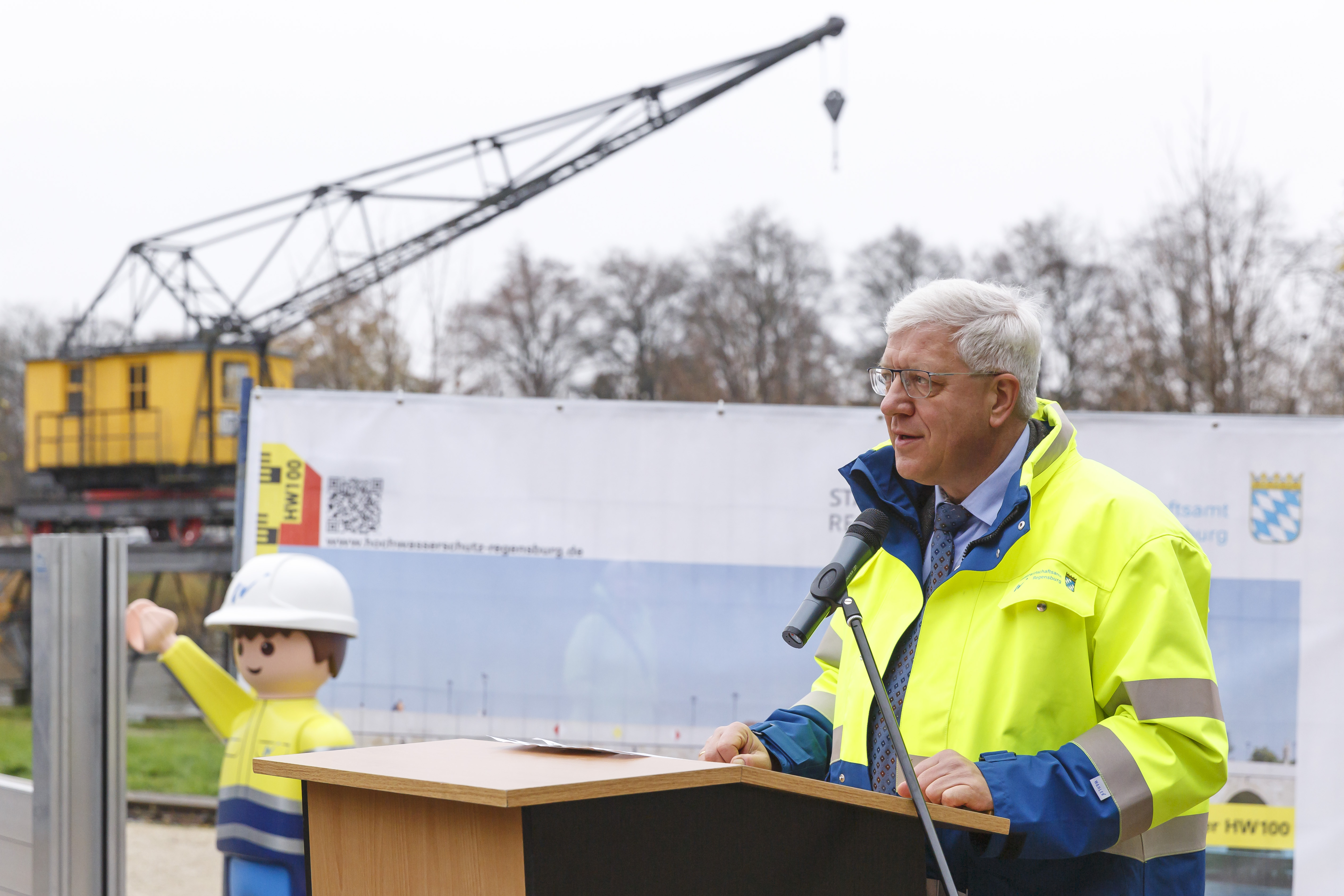 Rechts im Bild der Behördenleiter des WWAs am Rednerpult, dahinter ein alter, gelber Hafenkran, das Playmobilmännchen und ein Plakat zum Hochwasserschutz Regensburg.
