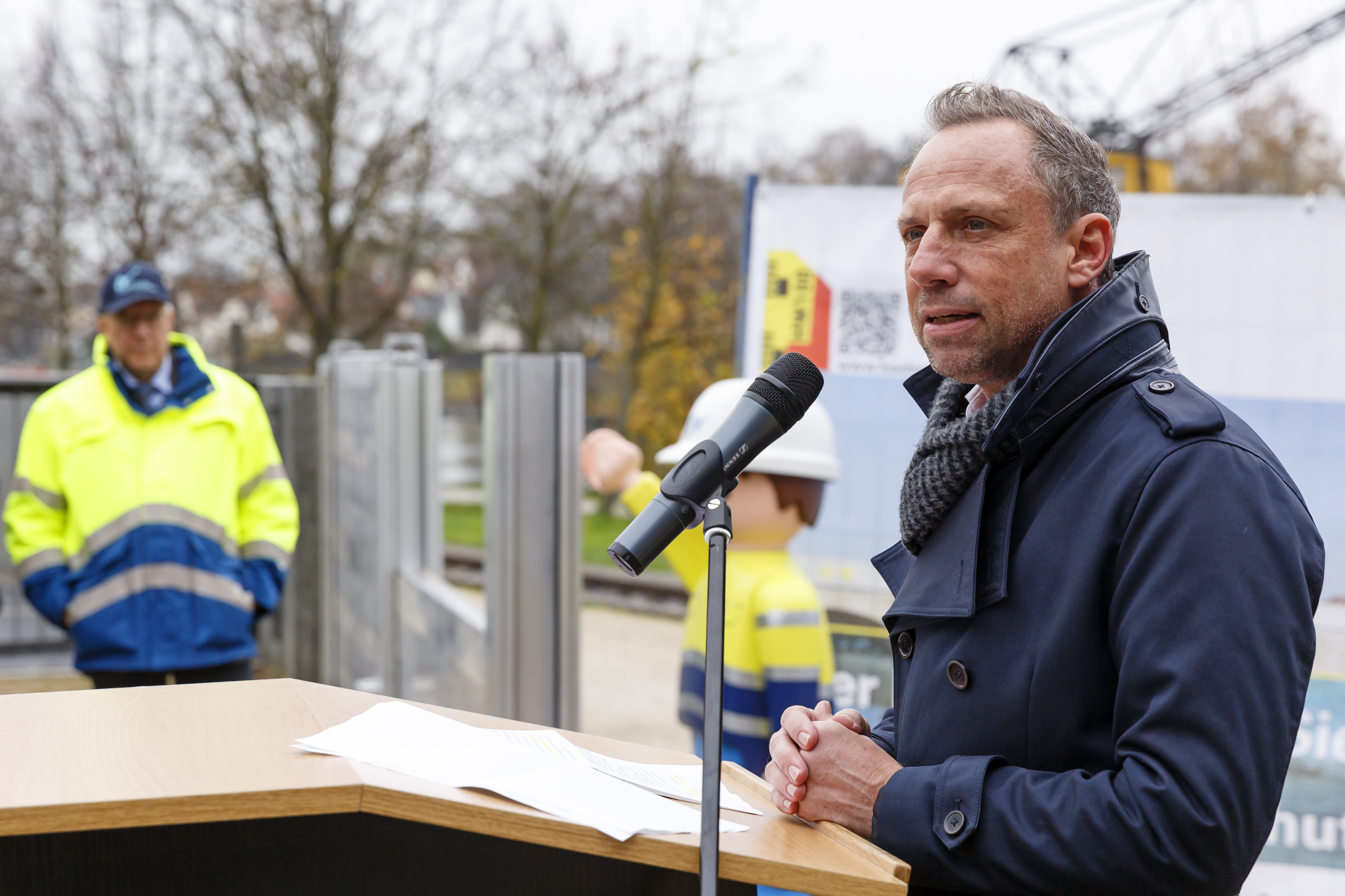 Der Minister steht am Rednerpult, im Hintergrund der Behördenleiter des WWAs, das Playmobilmännchen, die teilweise aufgebaute mobile Hochwasserschutzwand und ein Plakat zum Hochwasserschutz Regensburg.