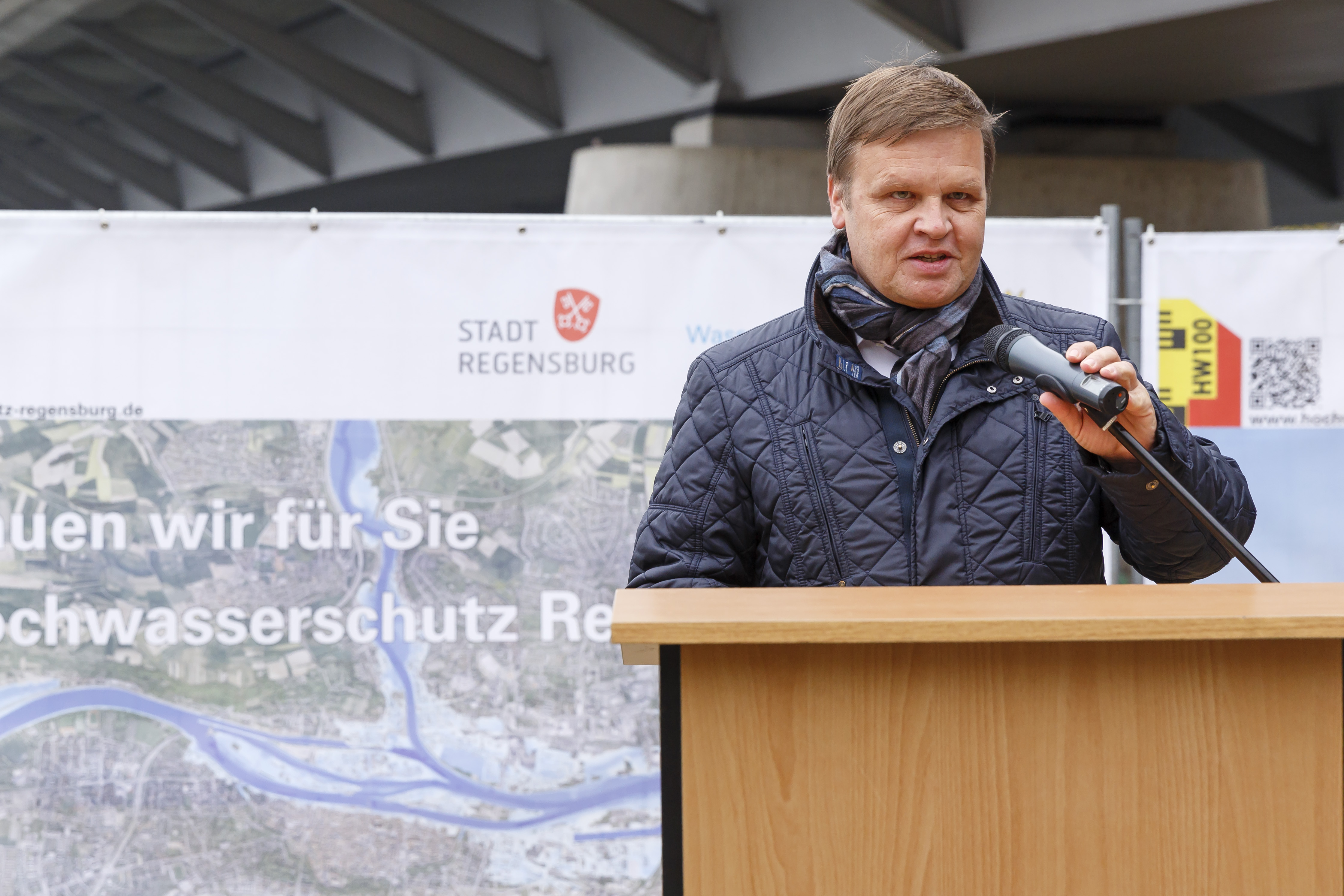 Der Geschäftsführer des Bayernhafen steht am Rednerpult, im Hintergrund ein Plakat zum Hochwasserschutz Regensburg und die Nibelungenbrücke.