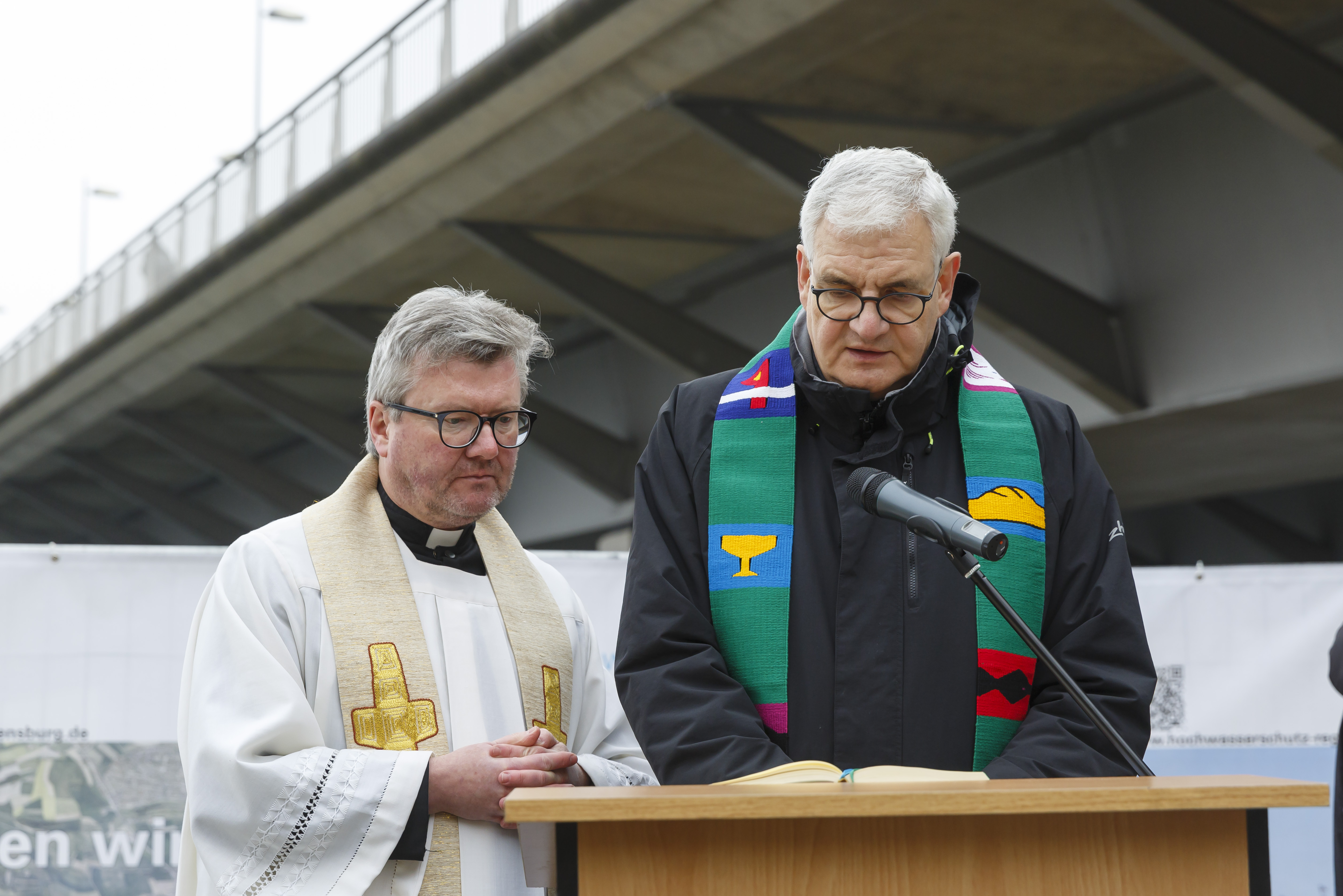 Ein katholischer und ein evangelischer Priester stehen am Rednerpult um den kirchlichen Segen zu spenden. Diagonal im Hintergrund die Nibelungenbrücke.