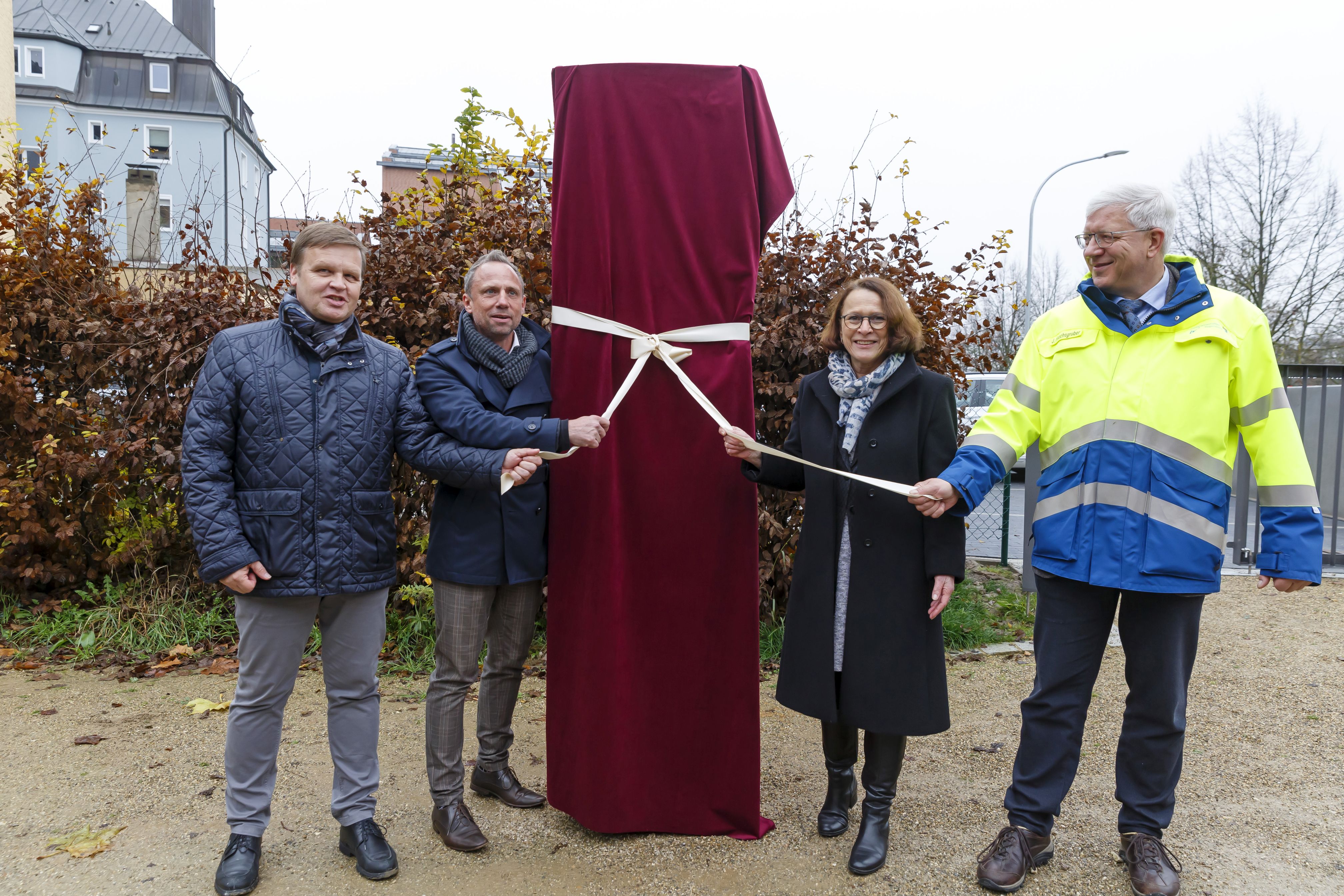 In der Mitte des Bildes die noch verhüllte Infostele, links daneben der Geschäftsführer des Bayernhafen und der Minister, rechts davon die Oberbürgermeisterin und der Behördenleiter des Wasserwirtschaftsamtes. Alle haben das Band zur Enthüllung der Stele in der Hand. Im Hintergrund Gebüsch und Bebauung.