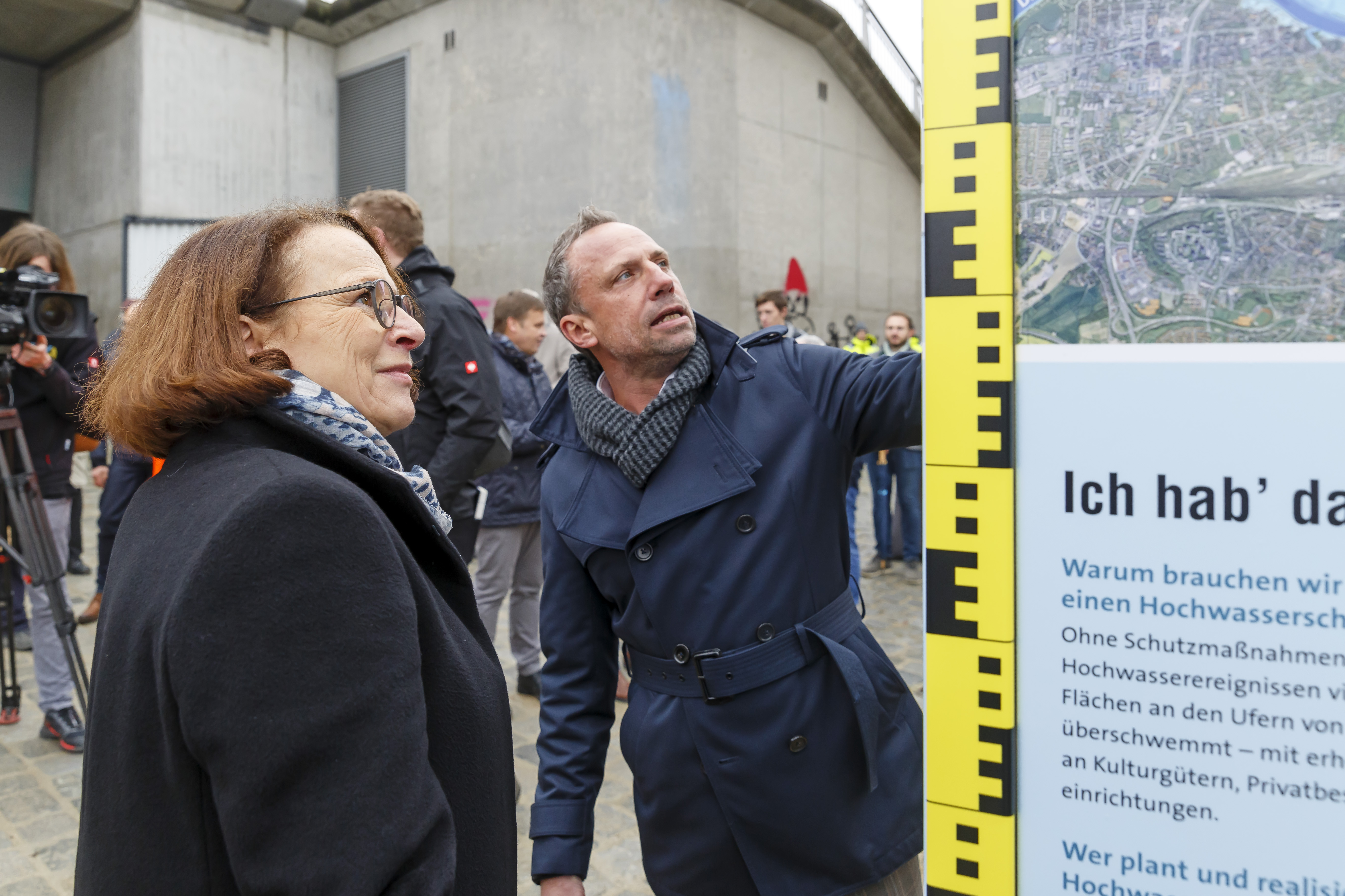 Die Oberbürgermeisterin und der Minister stehen interessiert an der rechts neben ihnen stehenden Infostele. Dahinter weitere Gäste der Einweihung. Im Hintergrund ist das Widerlager der Nibelungenbrücke zu sehen.