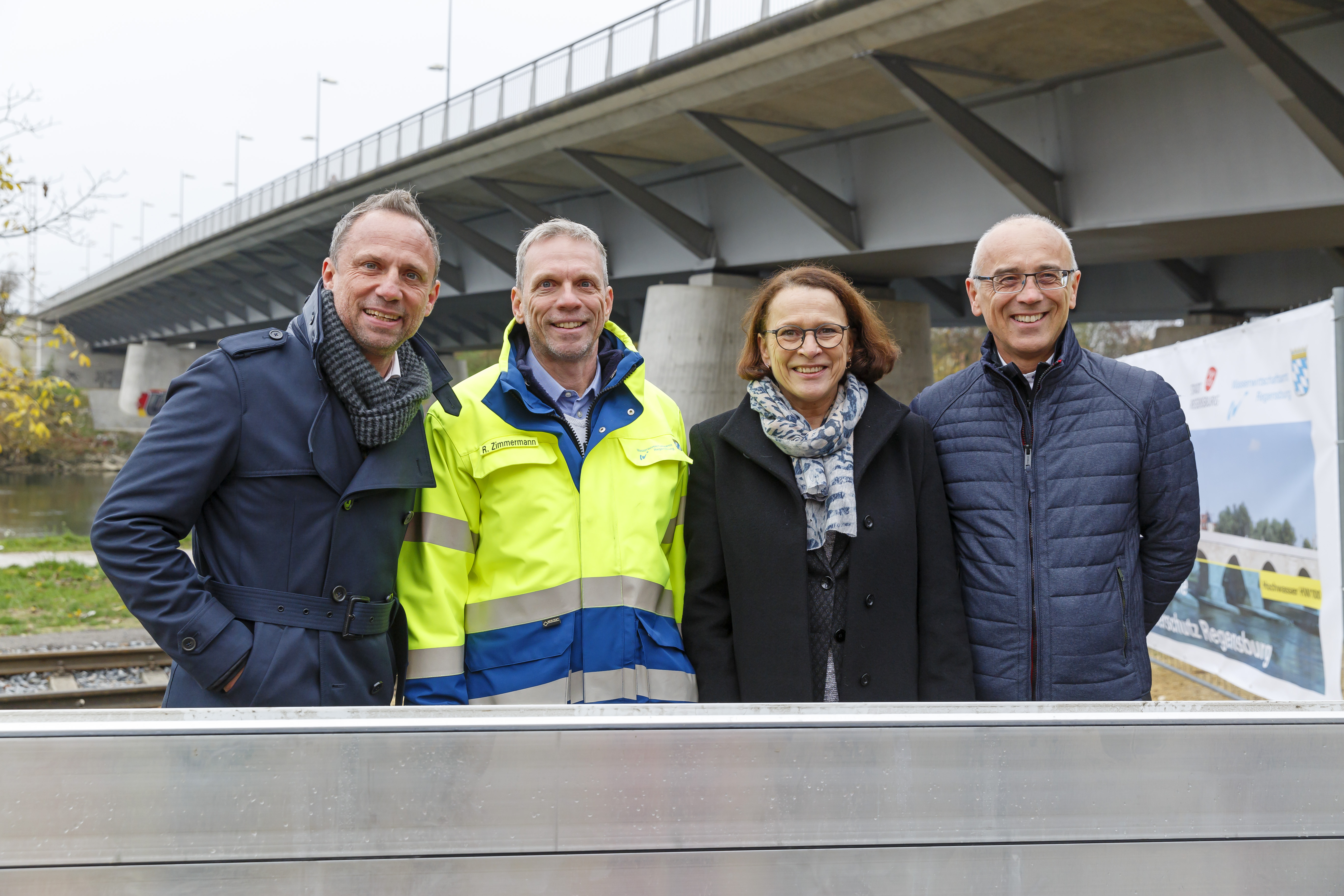 Im Vordergrund Dammbalken der mobilen Hochwasserschutzwand, dahinter der Minister, Projektleiter des WWA, Frau Oberbürgermeisterin und der Projektleiter der Stadt Regensburg. Im Hintergrund diagonal im Bild die Nibelungenbrücke.