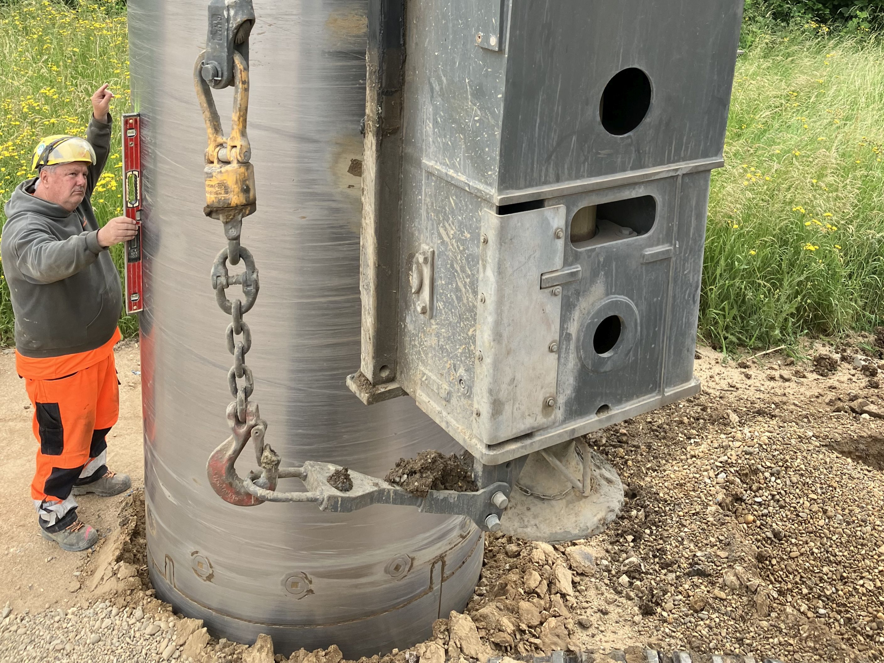 In der Mitte des Bildes hängt ein Rohr am Bohrgerät, das von einem links daneben stehenden Arbeiter mit der Wasserwaage in Position gebracht wird.