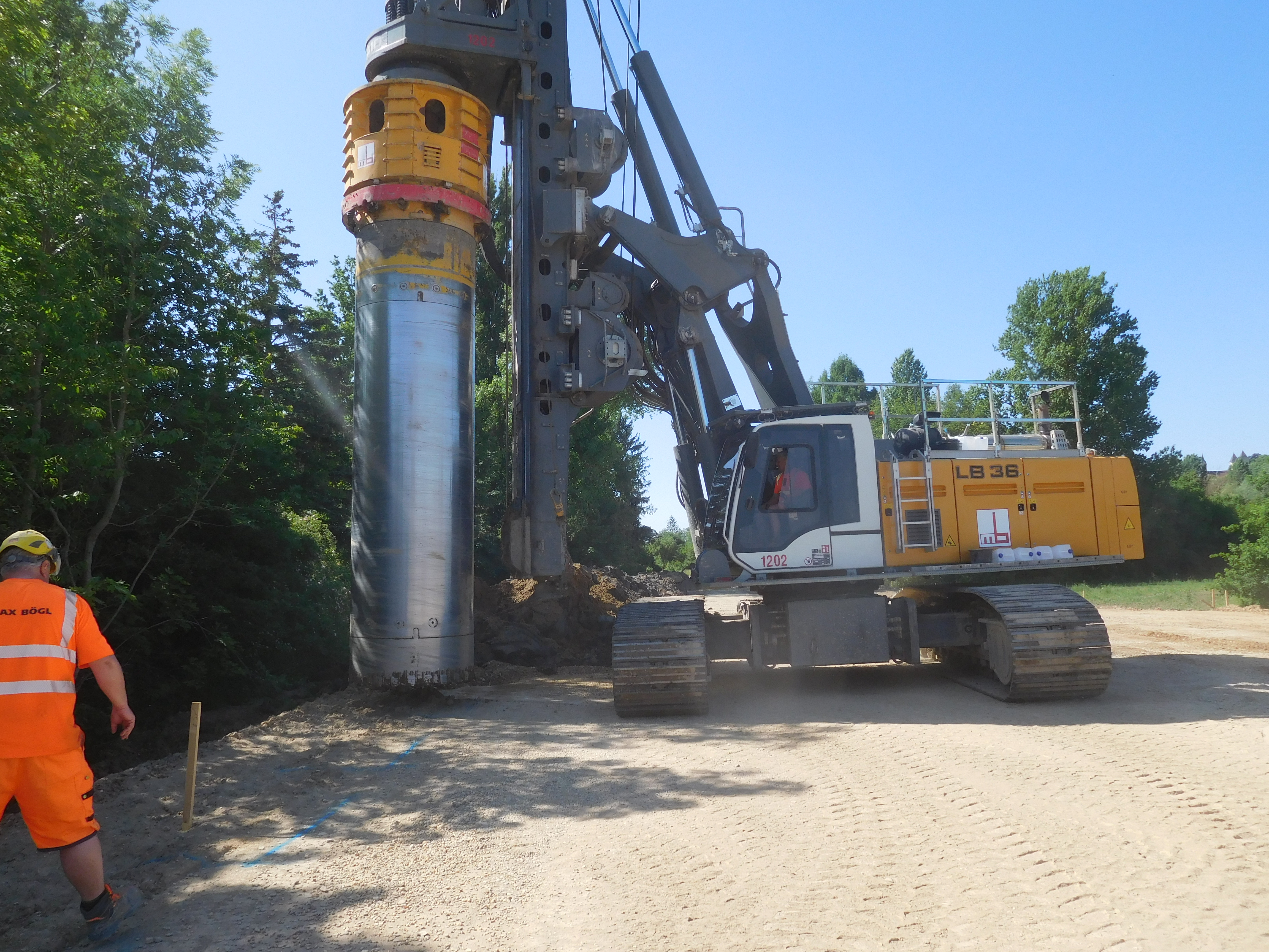 In der Mitte des Bildes ein Bohrgerät, der Bohrer wird in Position gebracht. Links daneben ein Bauarbeiter, Bäume im Hintergrund.