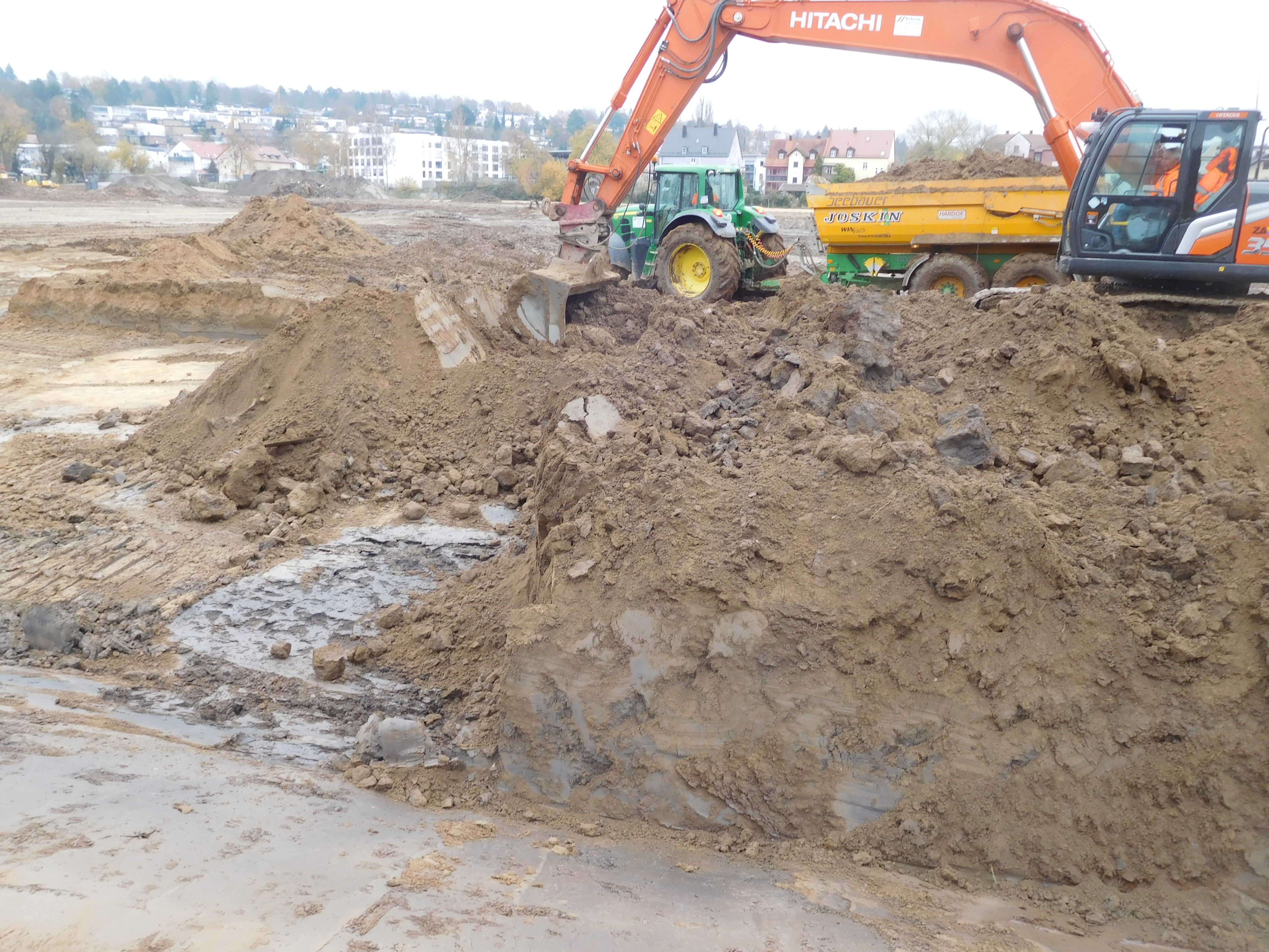 Ein Bagger steht auf einem Erdhaufen und lädt Erde auf einen im Hintergrund stehenden Traktoranhänger. Bebauung im Hintergrund.