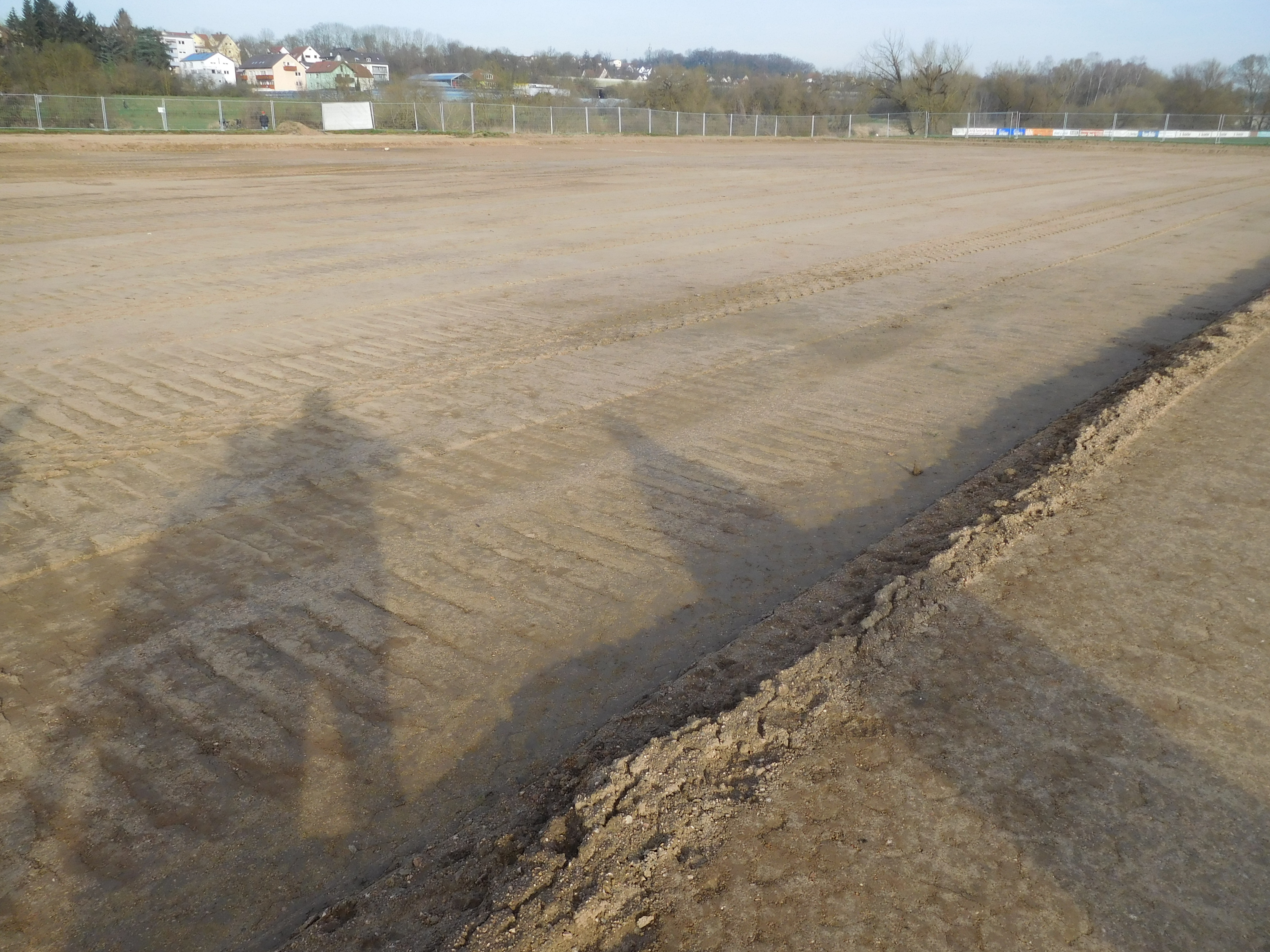 Die planierte Fläche des ehemaligen Sportplatzes. Der Bauzaun und Bebauung im Hintergrund.