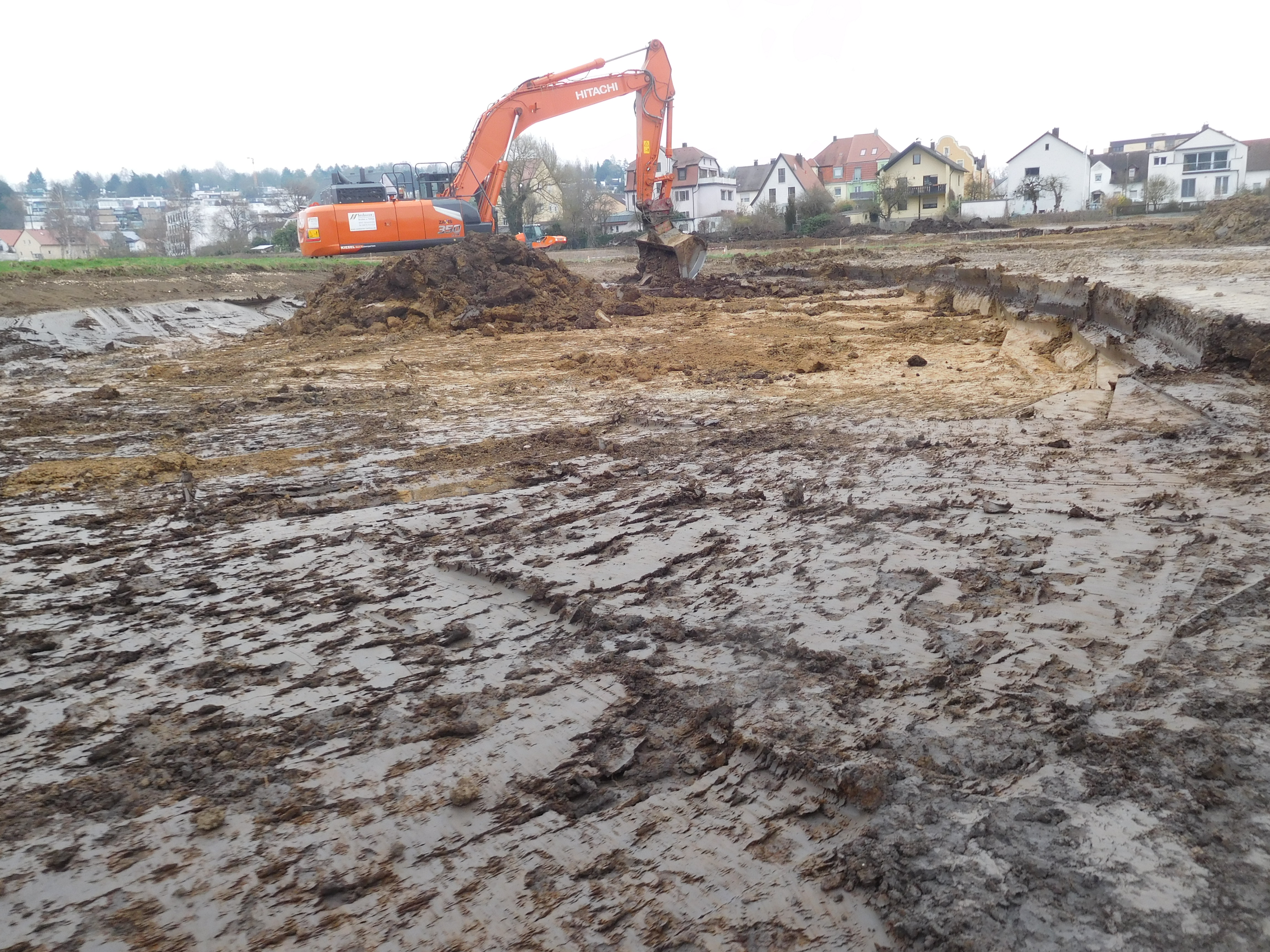 Ein Bagger in der Mitte des Bildes schiebt mit der Schaufel den Oberboden ab. Bebauung im Hintergrund.