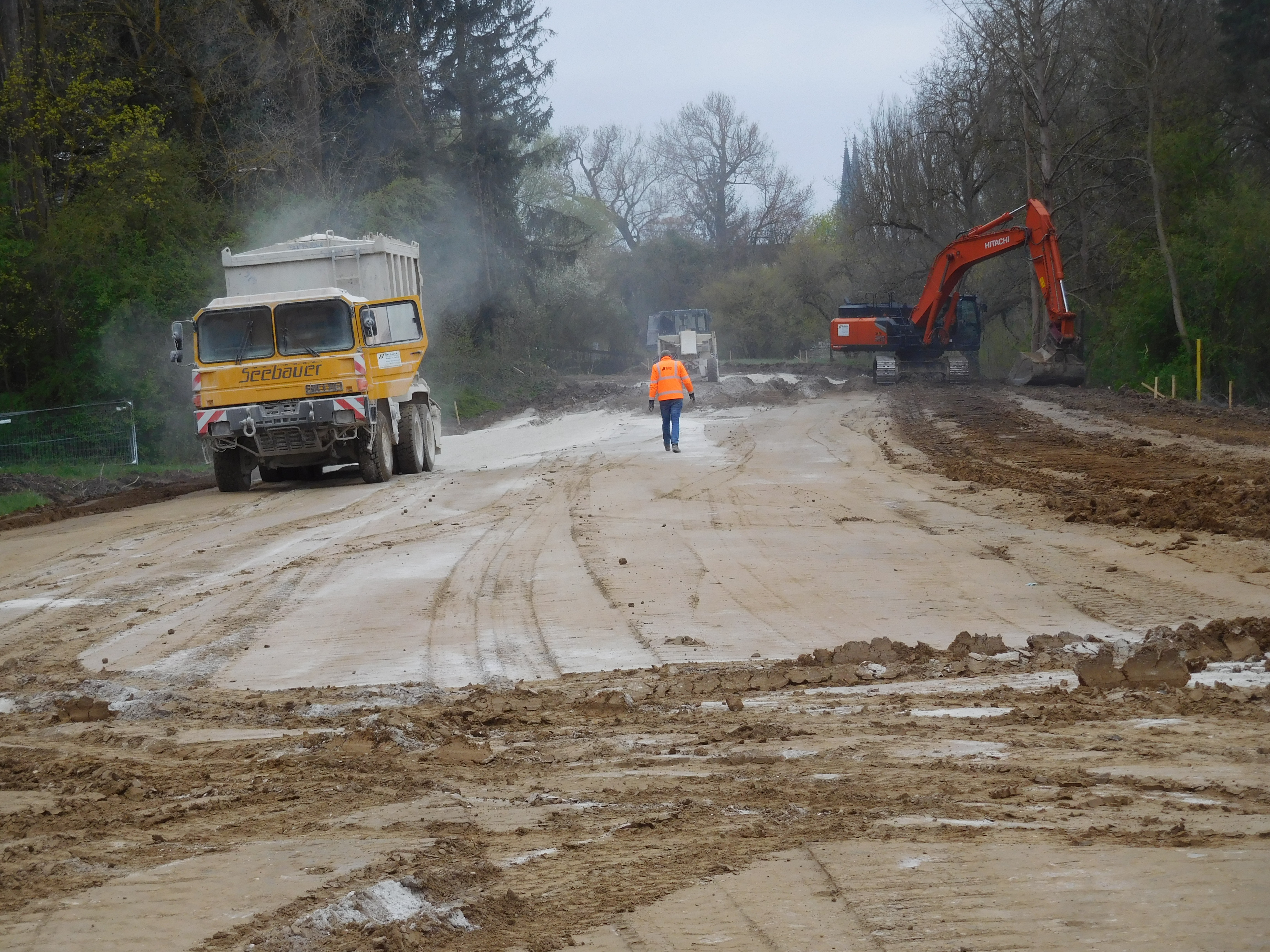 Auf der planierten Fläche links im Bild ein Lastwagen, rechts davon im Hintergrund eine Baumaschine und ein Bagger. Ein Bauarbeiter ist in der Mitte des Bildes von hinten zu sehen. Rund um die Baustelle sind Bäume und im Hintergrund sind die Domspitzen zu erkennen.