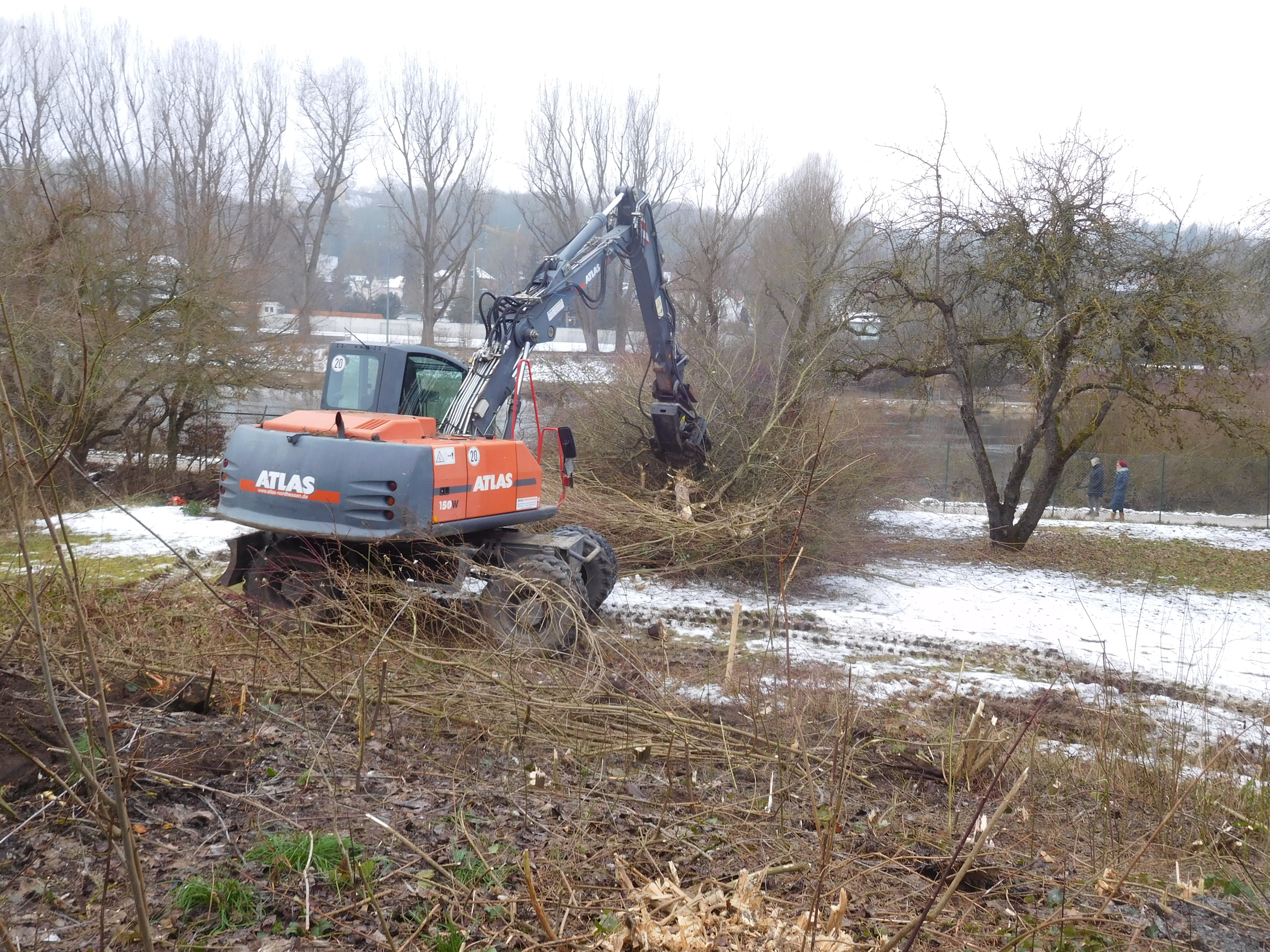 Eine Baumaschine mit Greifarm rodet das Gelände, im Hintergrund quert der Fluss Regen. Auf der anderen Uferseite ist ein Sportplatz zu sehen. Es liegt noch Schnee.