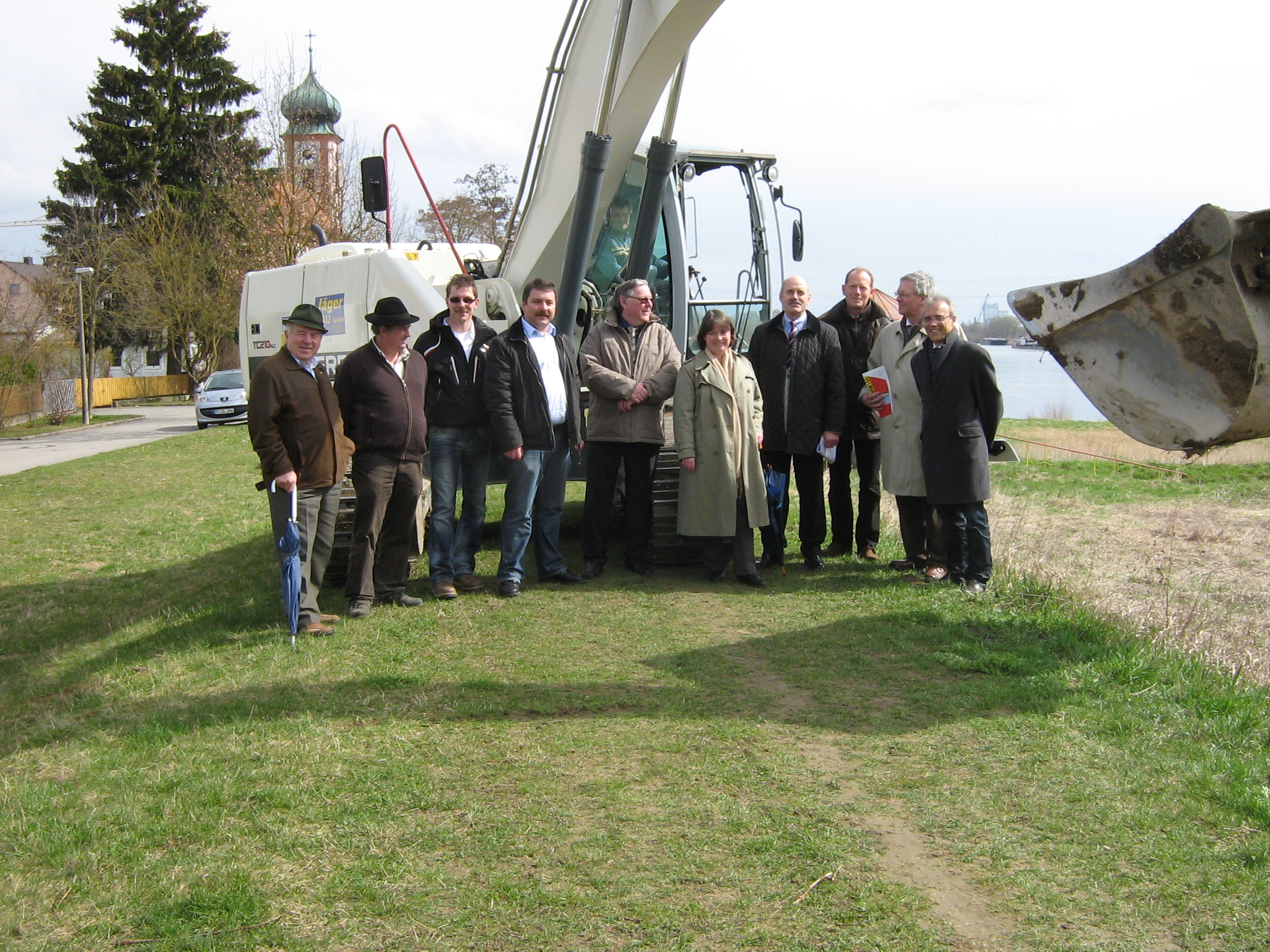 Spatenstich in Schwabelweis Oberbürgermeister Herr Schaidinger, Behördenleiter des WWA Regensburg, Herr Rogowsky und andere Vertreter der Stadt und Behörden, stehen vor einem großen Schaufelbagger am grünen Uferstreifen der Donau