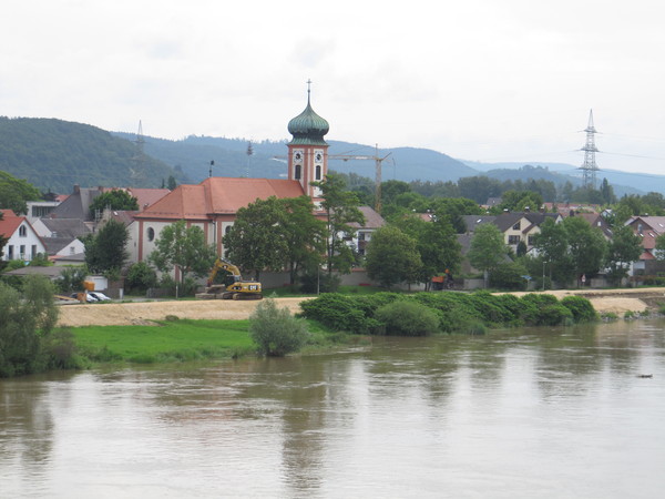 Donau mit Uferstreifen und Baustelle mit Bagger. Nahe am Ufer gebaute Häuser und die Kirche.