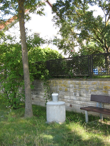 Eine Wiese mit einer Mauer, davor eine Bank, ein Baum und ein Grundwasserpegel.