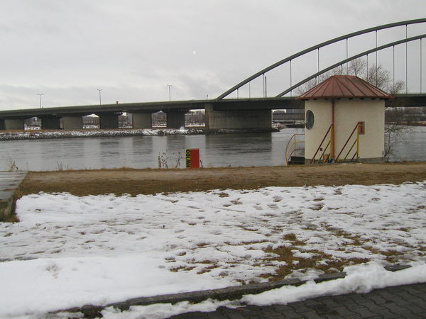Uferbereich mit Schnee und Pegelhaus an der Donau. Dahinter eine Brücke.