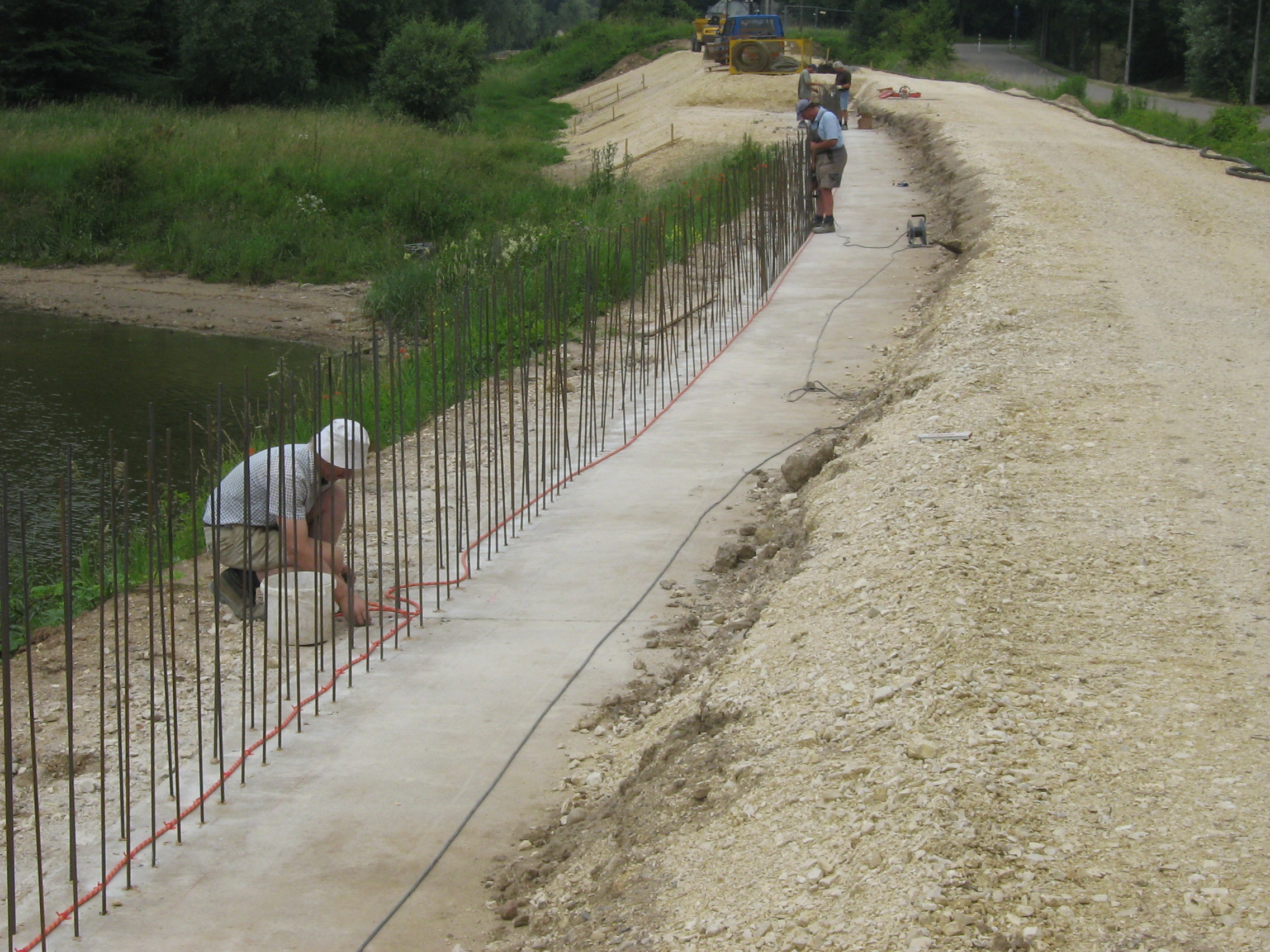 Diagonal im Bild wurden Baustahlstäbe einbetoniert. Zwei Bauarbeiter arbeiten dort, im Hintergrund weitere Bauarbeiter und Baumaschinen. Links die Böschung und das Donauufer, rechts davon der zukünftige Deichweg.