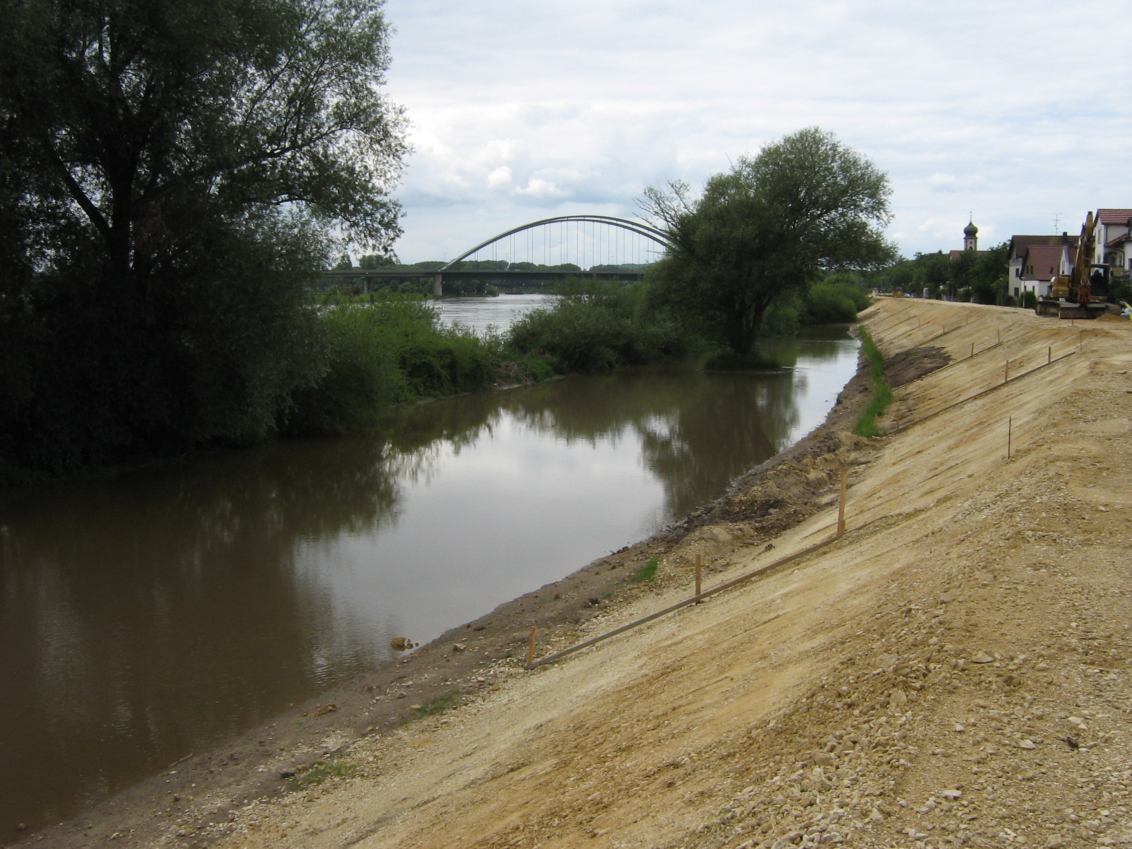 Rechts im Bild die aufgebaute Deichböschung, links davon die Donau teils mit Bewuchs. Im Hintergrund die Schwabelweiser Brücke, die Kirche und Häuser entlang des Deichs.