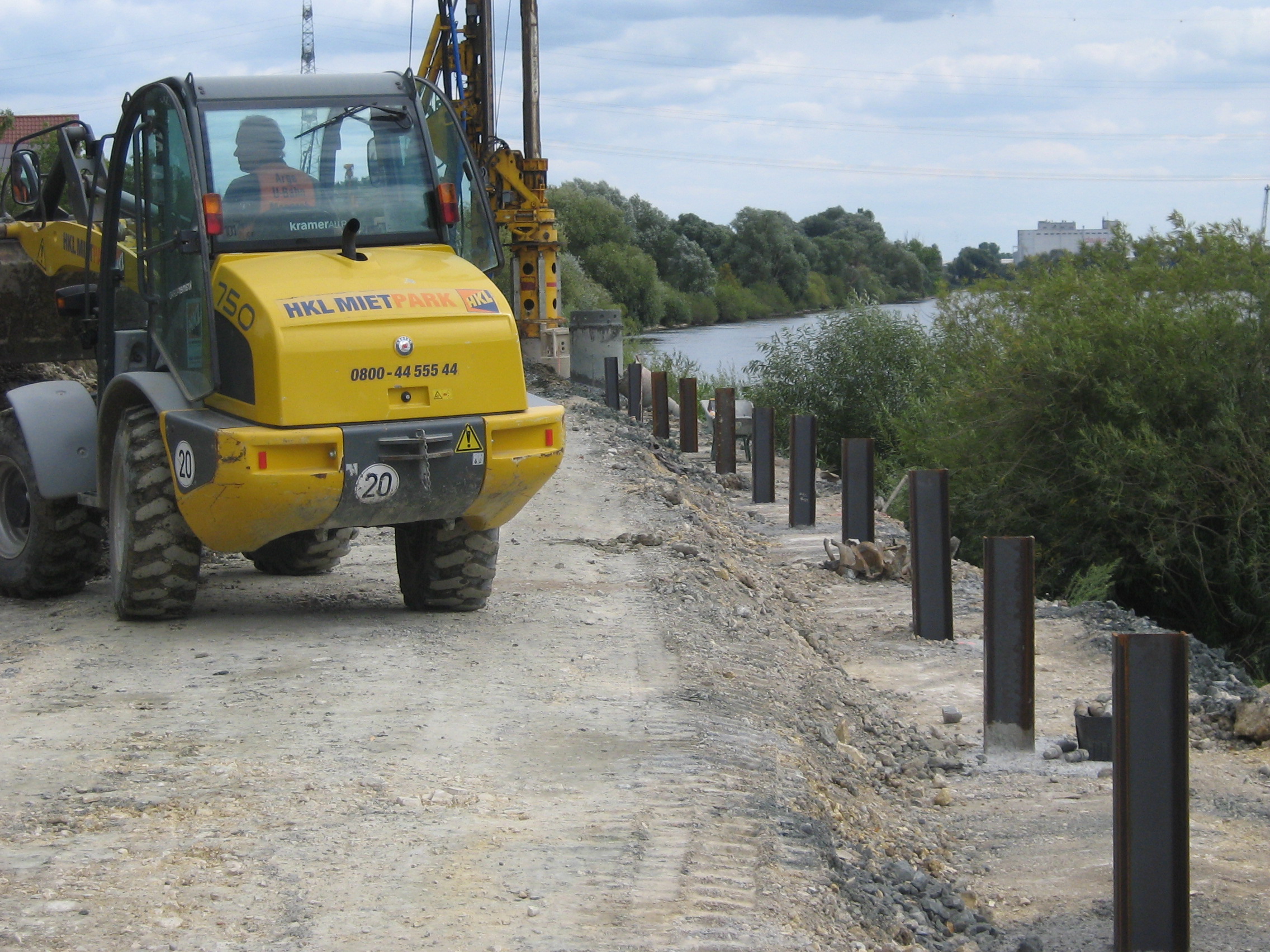 Links im Bild ein kleiner rückwärts fahrender Bagger, dahinter eine Art Bohrer der die Stahlträger mit Beton in den Boden einbringt, rechts eine Reihe schon eingebrachter Stahlträger, die Uferböschung und die Donau