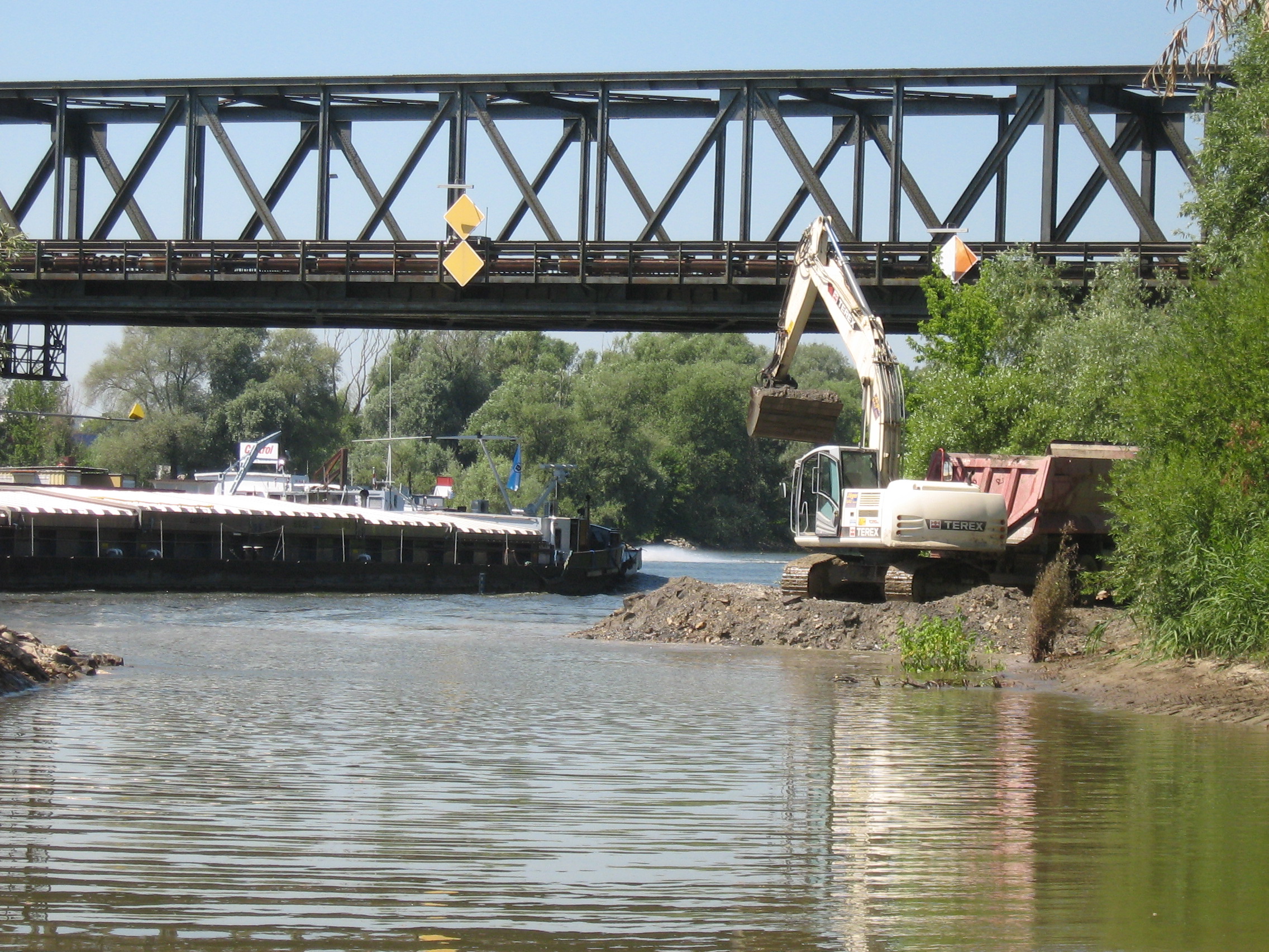 Der Blick aus dem Nebenarm in die Donau. Rechts im Bild ein grabender Bagger der einen LKW belädt. Im Hintergrund auf der Donau ein Frachtschiff, darüber quert die Eisenbahnbrücke.