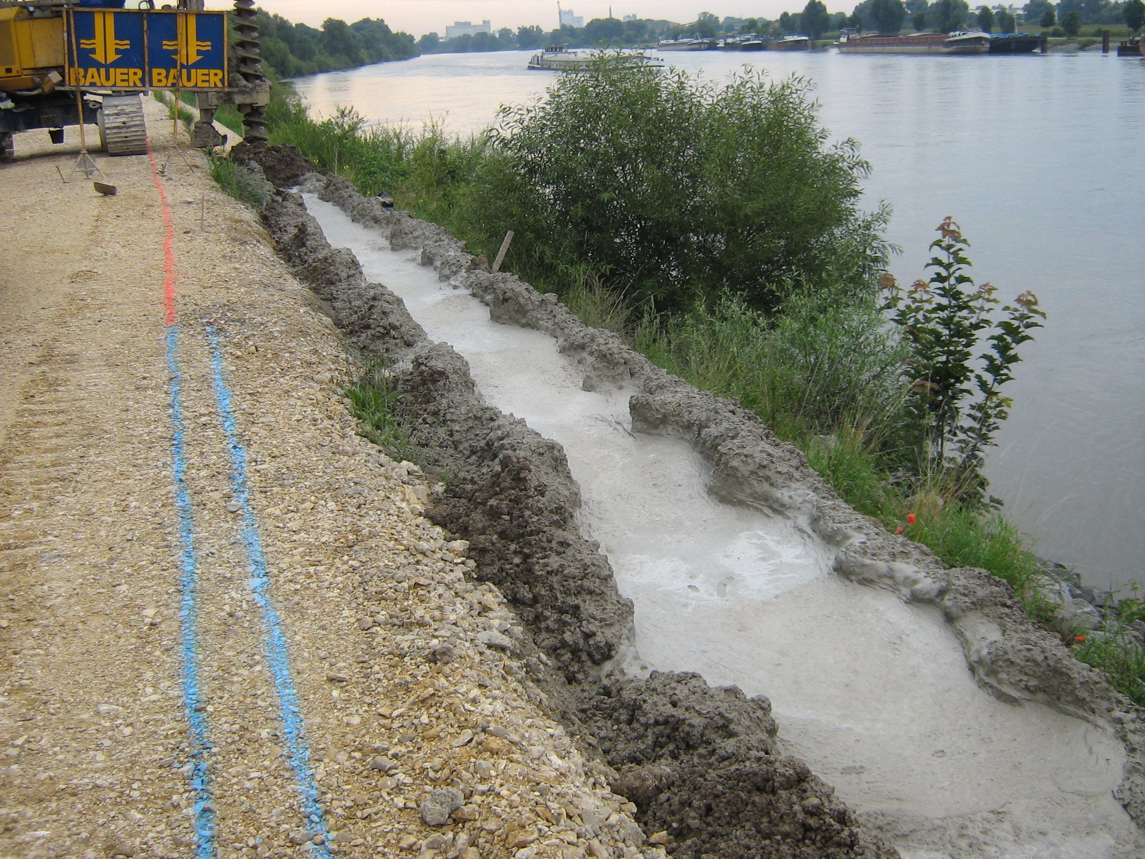 In der Mitte des Bildes wird die MIP Wand eingebracht. Bei diesem Verfahren, wird gebohrt und zugleich der Beton eingebracht. Im Hintergrund sieht man die Baumaschine mit dem Bohrer. Links davon das Gelände und rechts die Böschung, die Donau und einige Frachtschiffe.