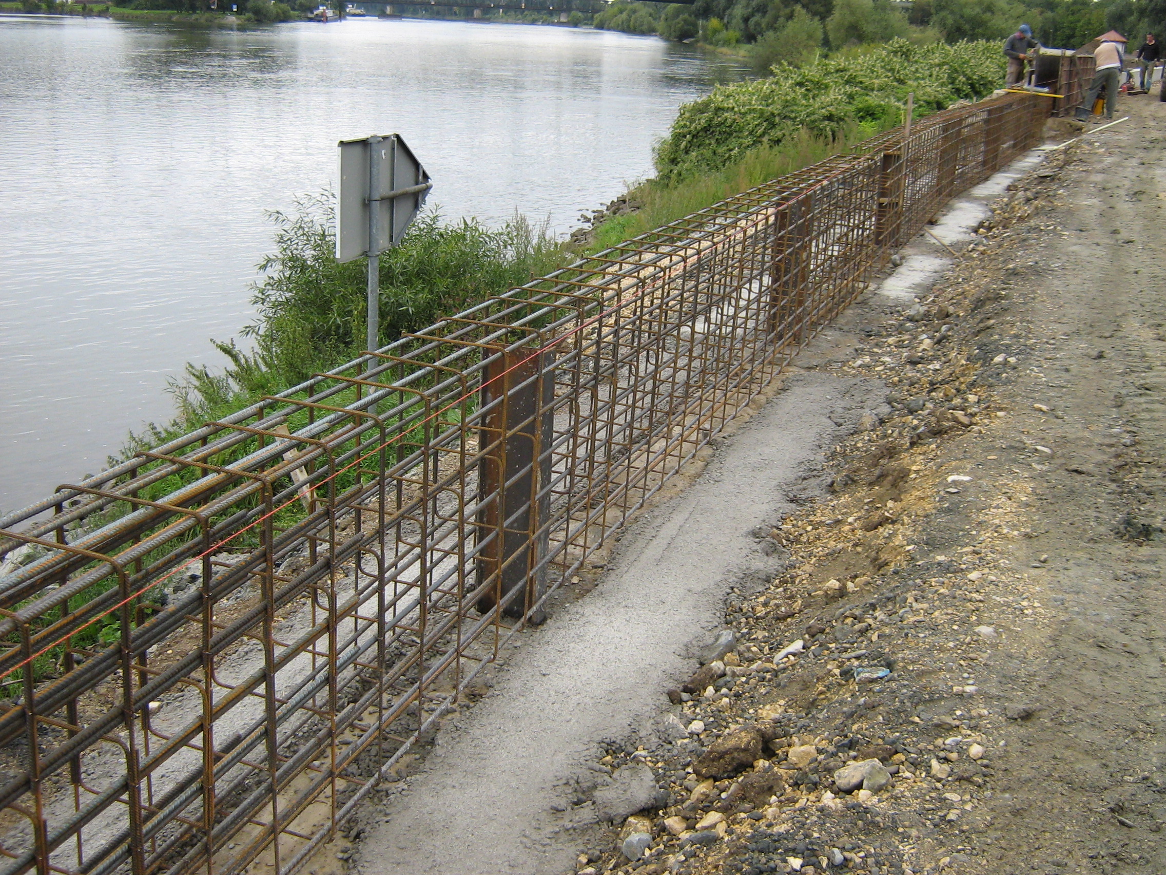 diagonal im Bild ein einbetoniertes Stahldrahtgeflecht in der Form einer Mauer, dahinter die Donau mit begrünter Böschung, rechts im Bild Füllmaterial, im Hintergrund Bauarbeiter