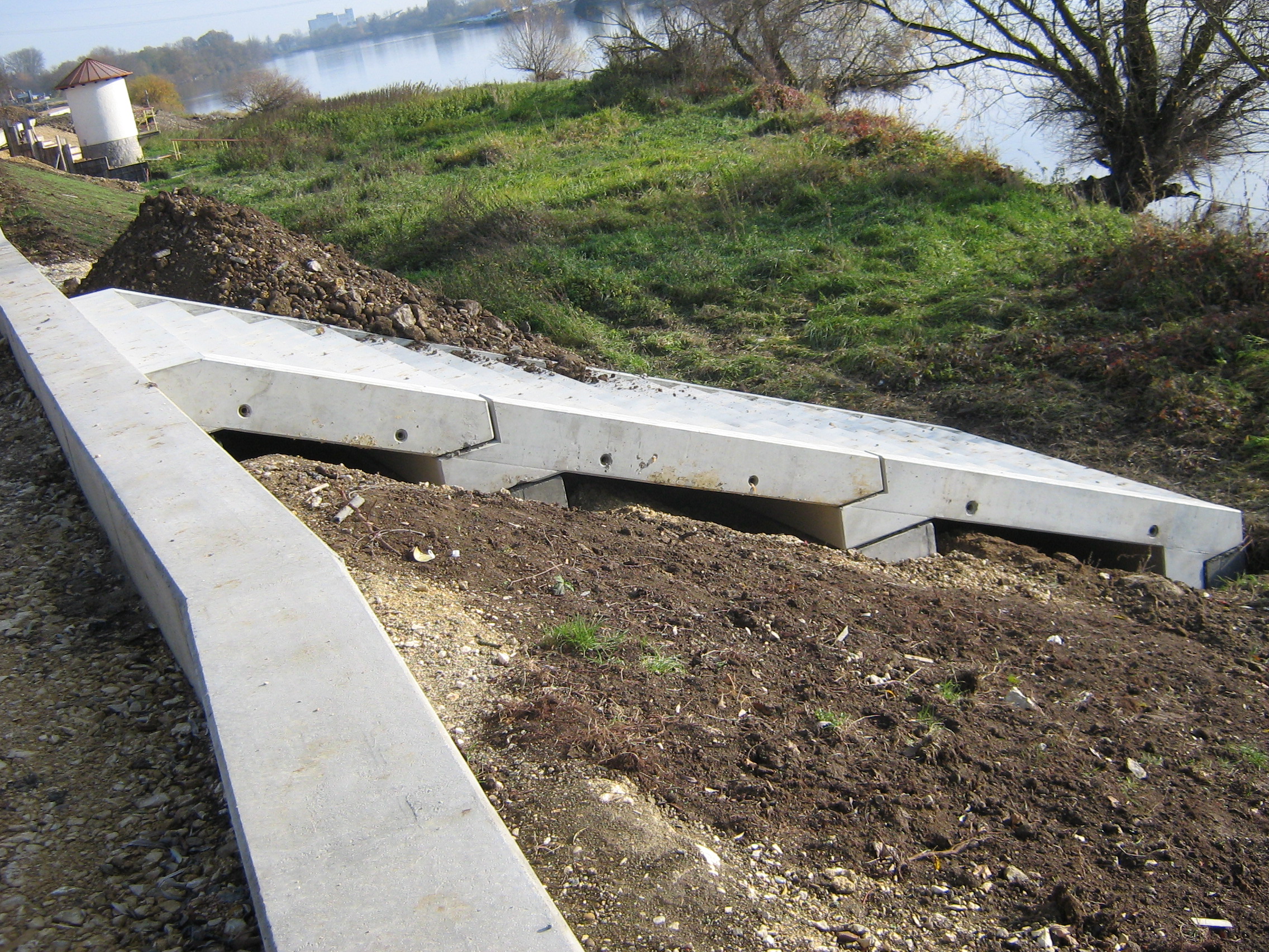 Links entlang des Bildes verläuft der Kopfbalken, quer dazu die Treppe an der Böschung aus zusammengesetzten Betonfertigteilen. Im Hintergrund das Pegelhaus, das begrünte Ufer und die Donau.