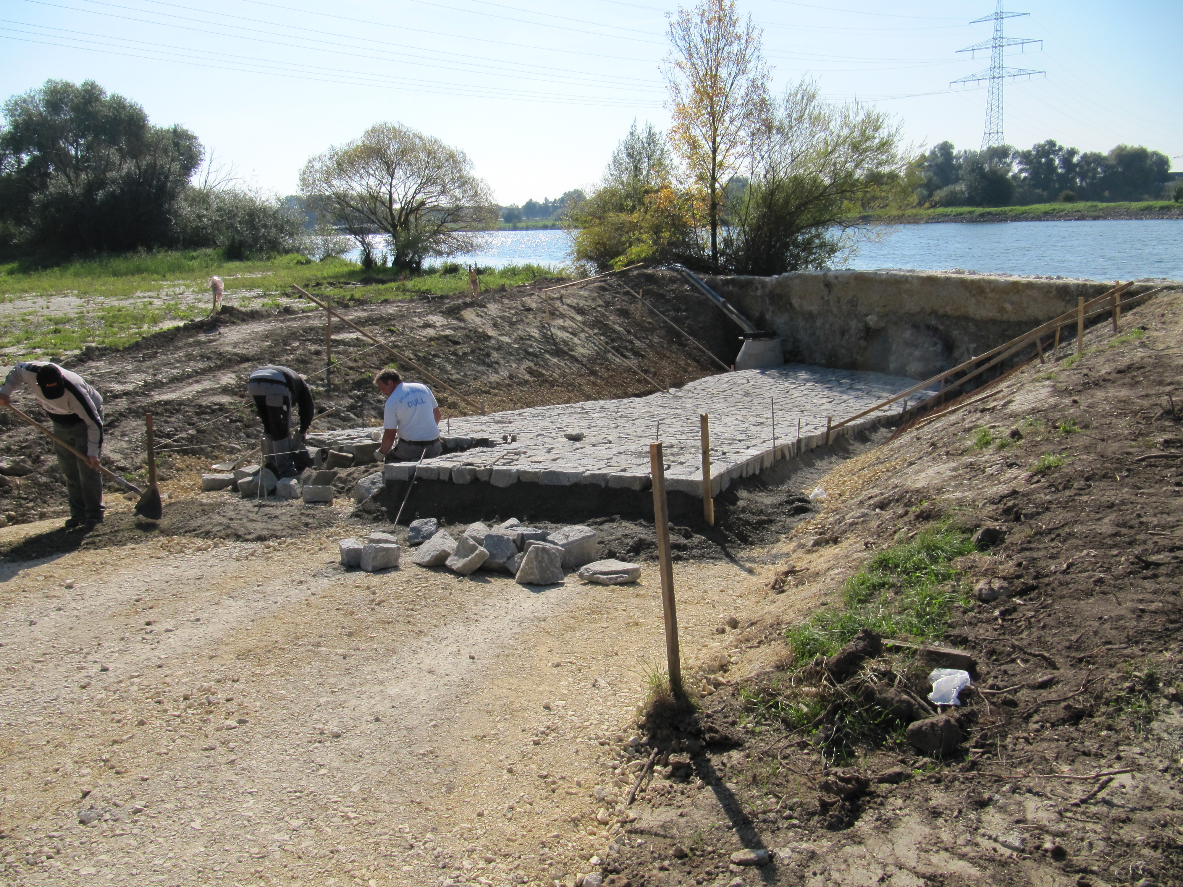 Zentral im Bild sind drei Bauarbeiter beim Pflastern der Slipanlage. Im Hintergrund quert die Donau und das begrünte Donauufer.