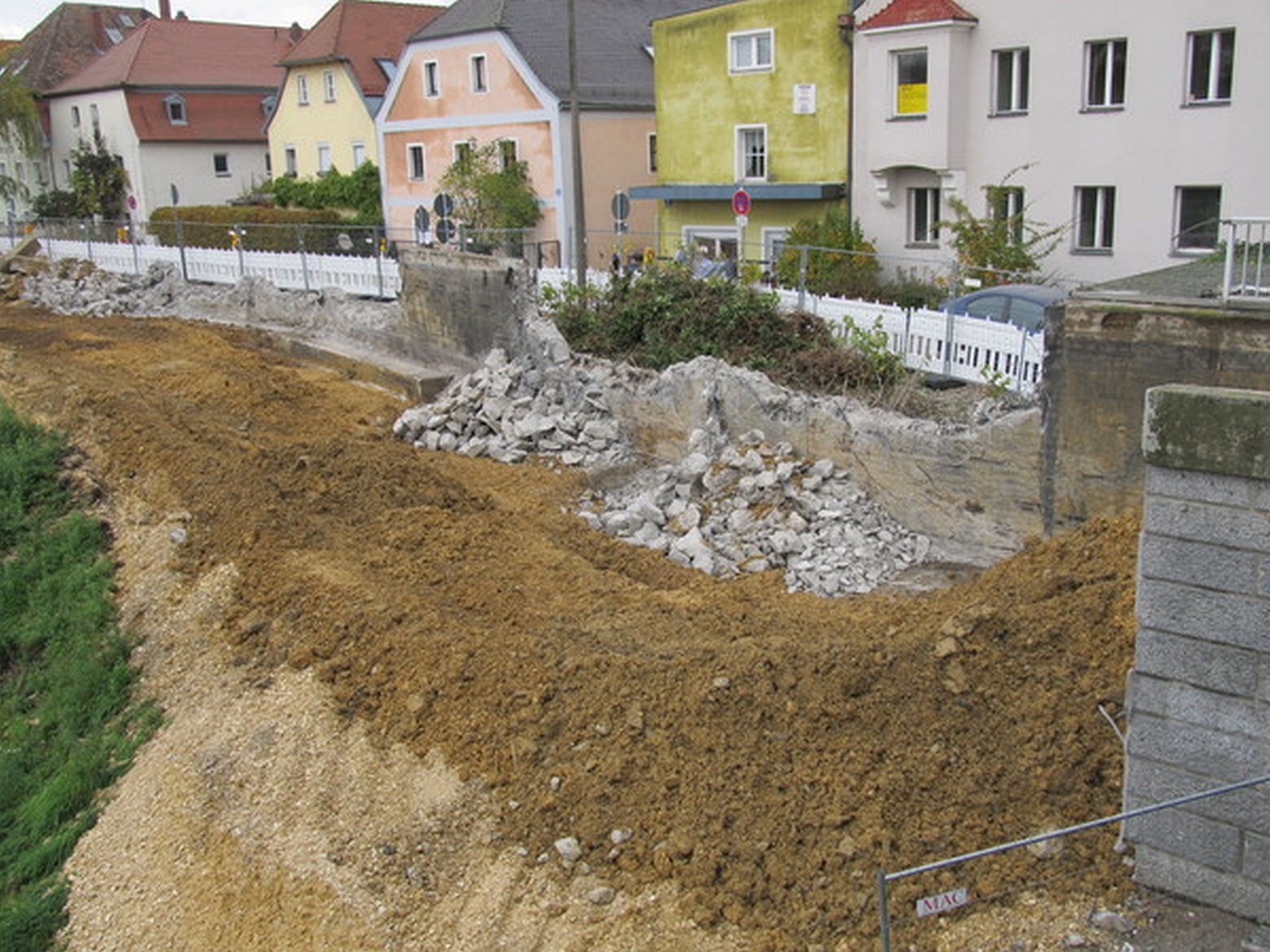 Im Vordergrund liegt Erdreich, Sand und Schotter, dahinter die bereits teilweise abgebrochene Betonwand. Im Hintergrund Bauzäune und die farbige Häuserreihe der Oberen Regenstraße. Rechts im Bild das Widerlager der Reinhausener Brücke.